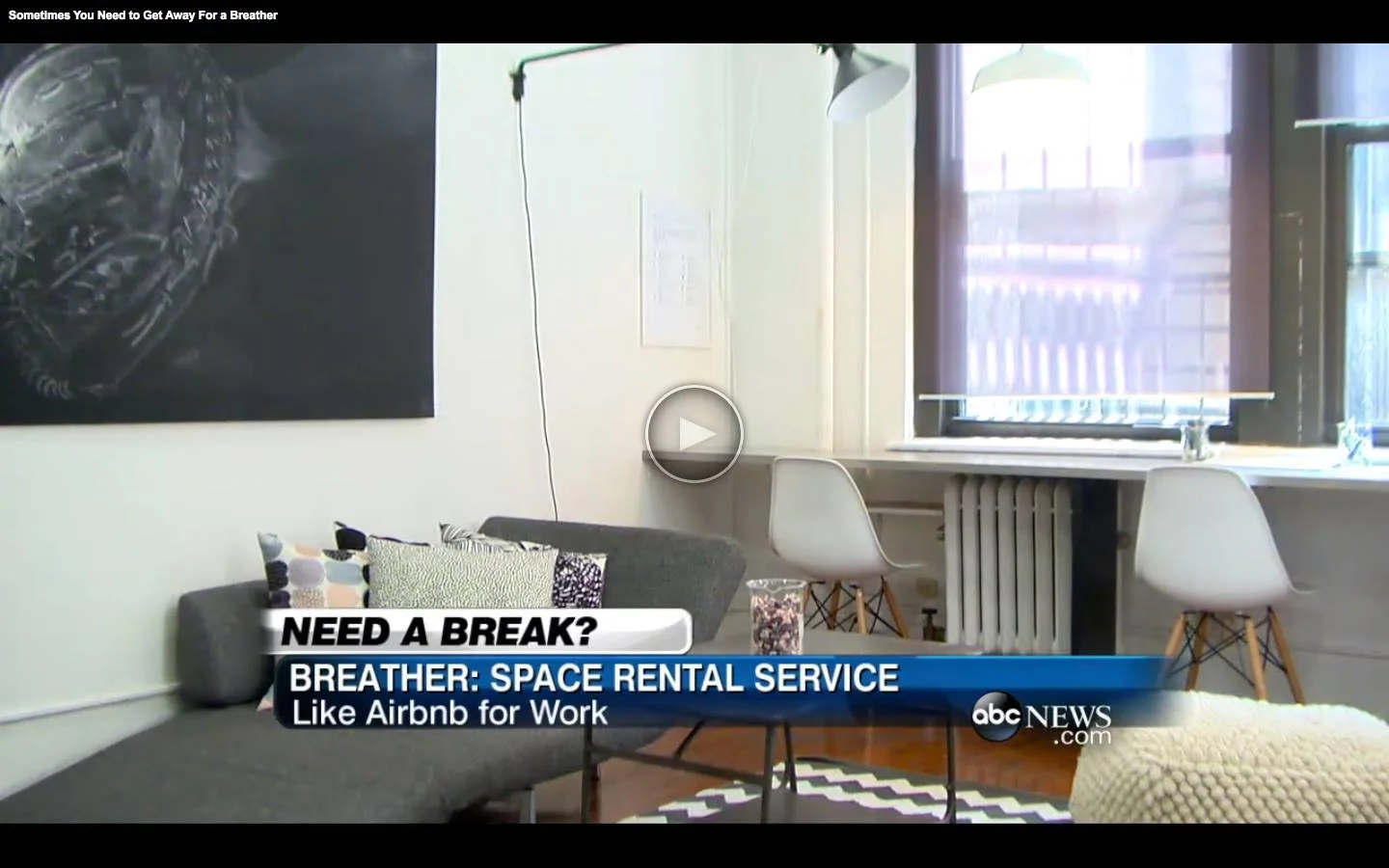 Living room with a gray sofa, pillows, a small round black table, a white desk with two white chairs, a large window with blinds, and a radiator. Text overlay about space rental service.