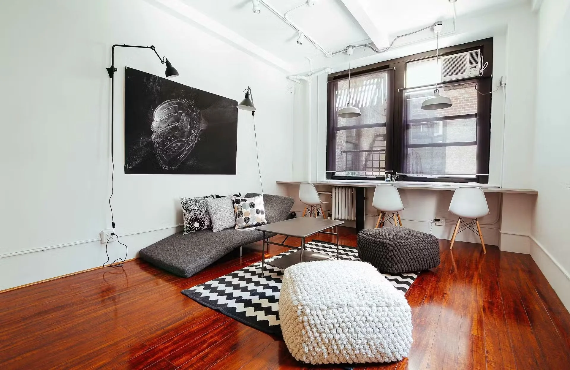 A modern living room with hardwood floors, a gray sectional sofa with patterned pillows, and a black and white chevron rug. There are two textured ottomans, one white and one dark gray. The wall above the sofa features a large black artwork, and two wall-mounted reading lamps. Large windows with dark frames and blinds let in natural light, and there are two white hanging pendant lights. An air conditioning unit is installed in the window.