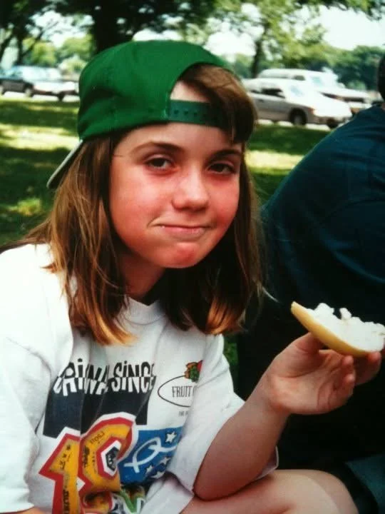 A young girl with shoulder-length brown hair wearing a green baseball cap backward, sitting outdoors on a sunny day, holding a bowl with food, and smirking at the camera.