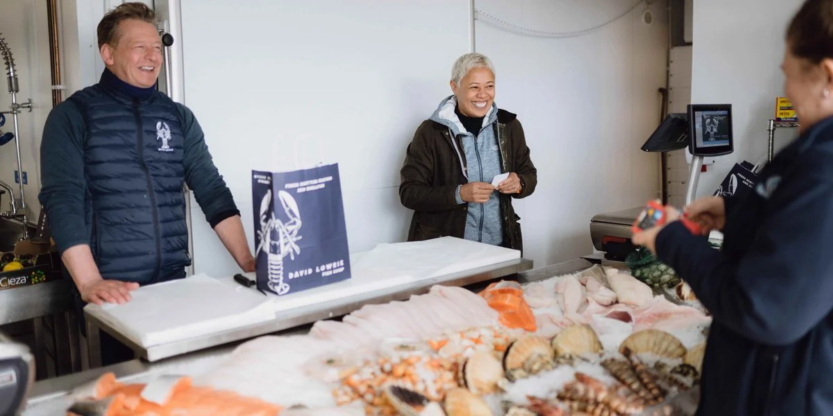 A seafood market with two men and one woman, all smiling. The woman and one man are behind the counter, and the woman is holding a small item while the other man is looking at her. The counter has various seafood, including lobsters and fish, displayed on ice.