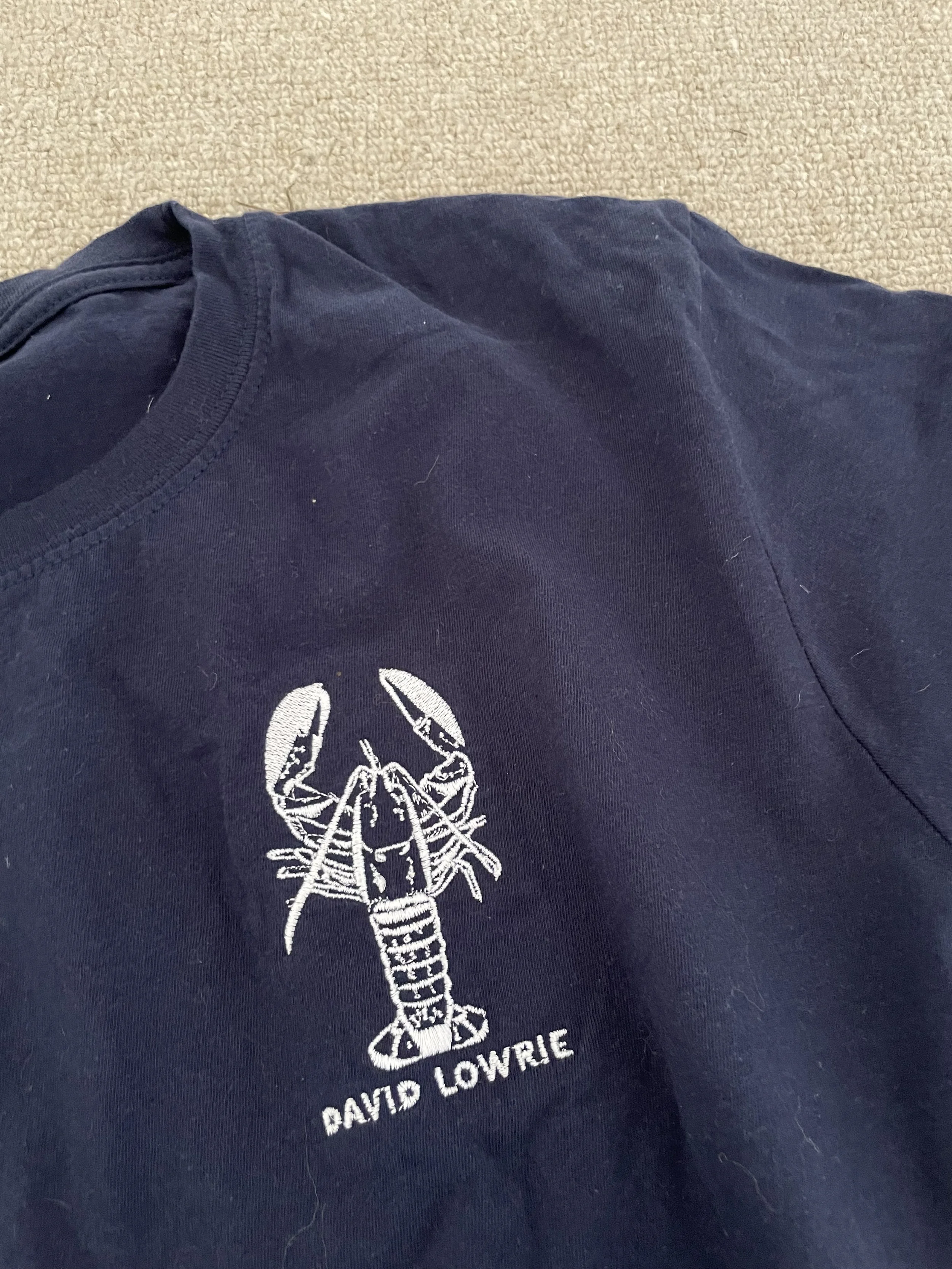 Close-up of a navy blue T-shirt with a lobster illustration and the text 'David Lowrie' embroidered in white.