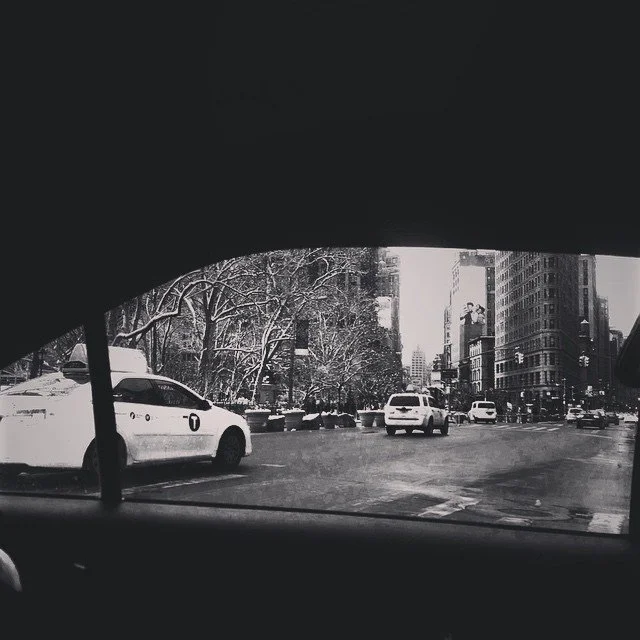City street view seen from inside a vehicle, with snow-covered trees, parked cars, and tall buildings.