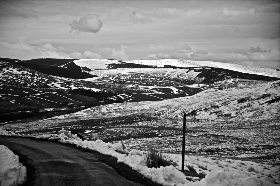 Snow-covered rolling hills and mountains, with a winding road in the foreground, under a cloudy sky, in a rural landscape during winter.