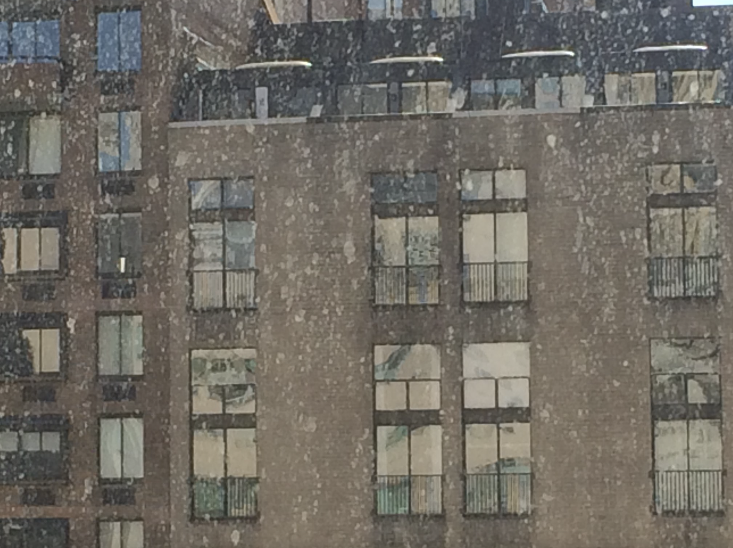 A tall brick apartment building with multiple windows and balconies, some with laundry hanging. Reflections of the surrounding cityscape are seen in the windows.