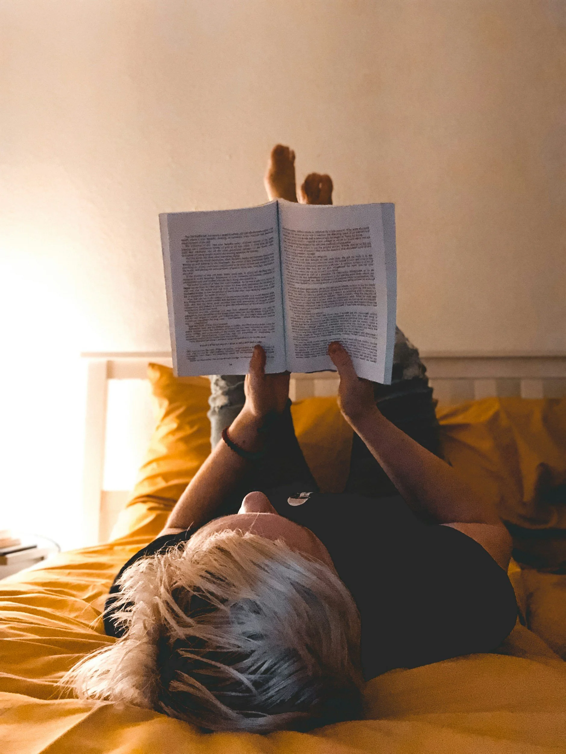 A person with blonde hair lying on a bed with yellow sheets, reading a book while a small dog with white and brown fur rests beside them.