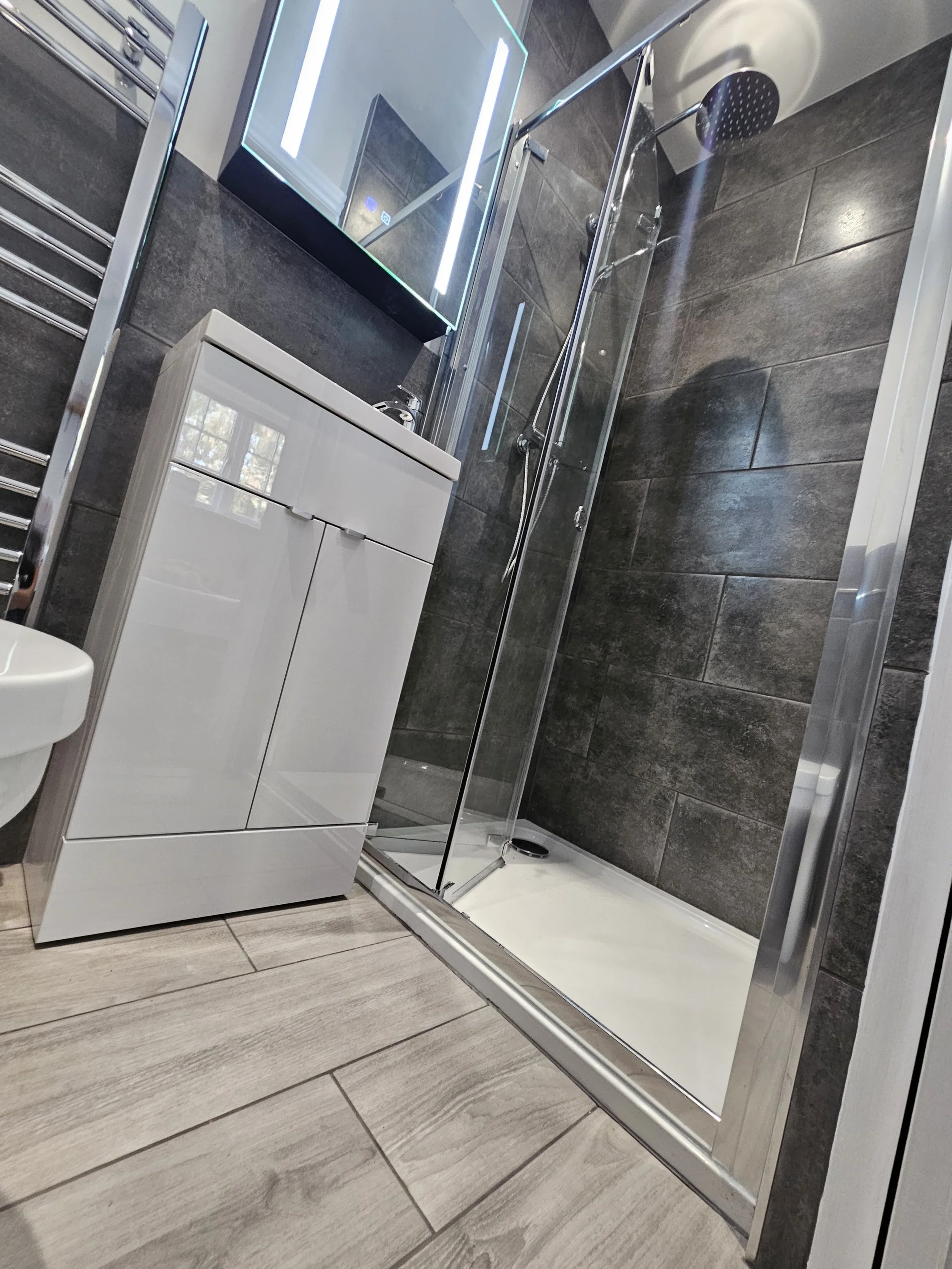 A modern bathroom shower area with dark gray tiles, a clear glass shower door, a white vanity cabinet, and a large LED mirror with backlit lighting.