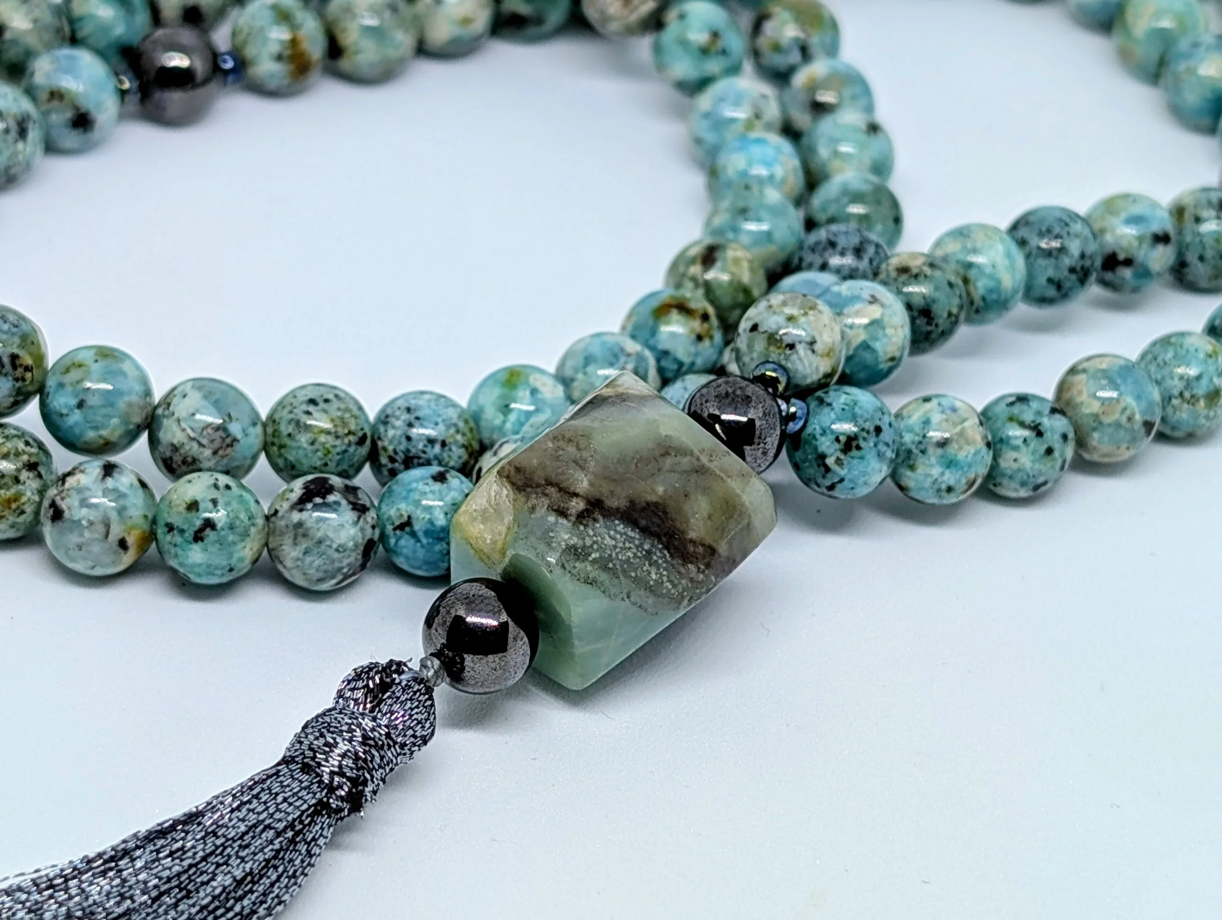 Close-up of turquoise and black beaded necklace with a large stone pendant, held by a person's hand with black-and-white patterned bracelet, on a white background.