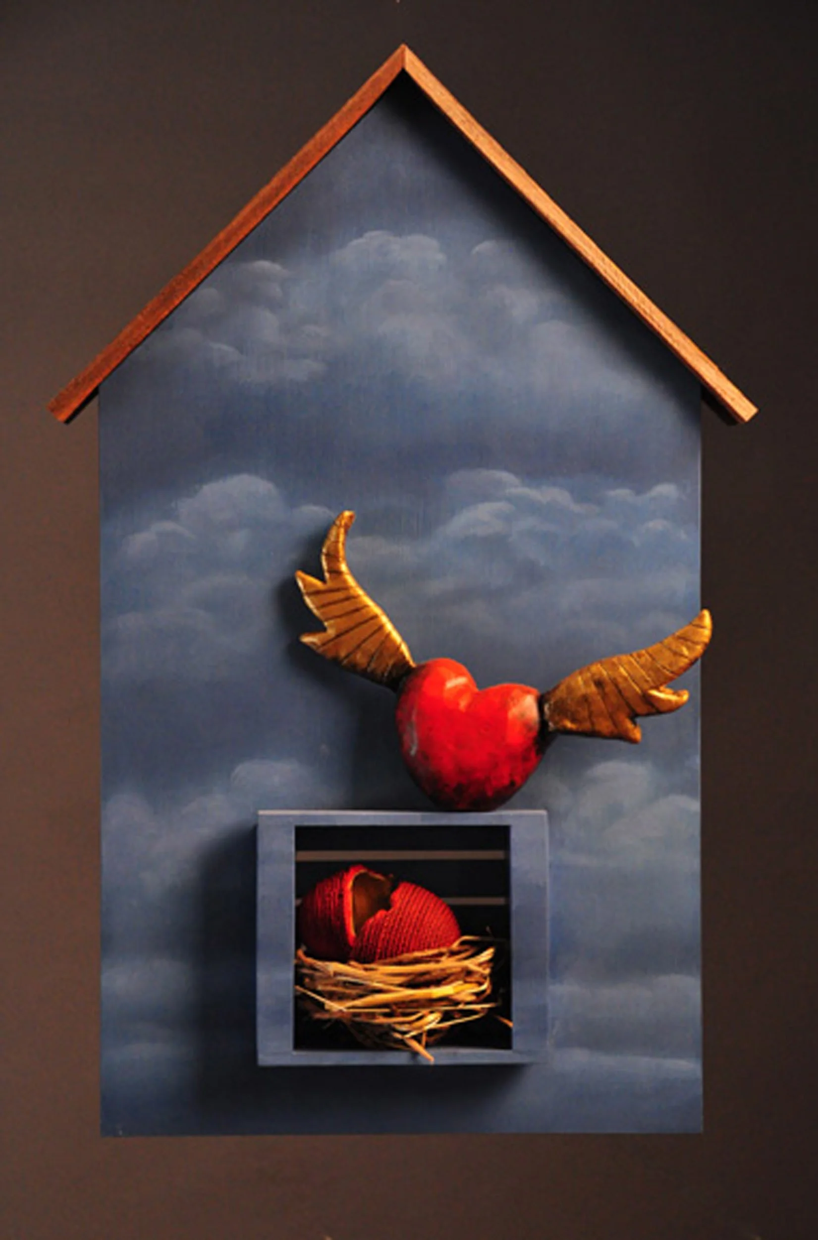 A miniature house with a painted sky background, featuring a wooden roof, a decorative wind, and a small basket with a red knitted item inside.