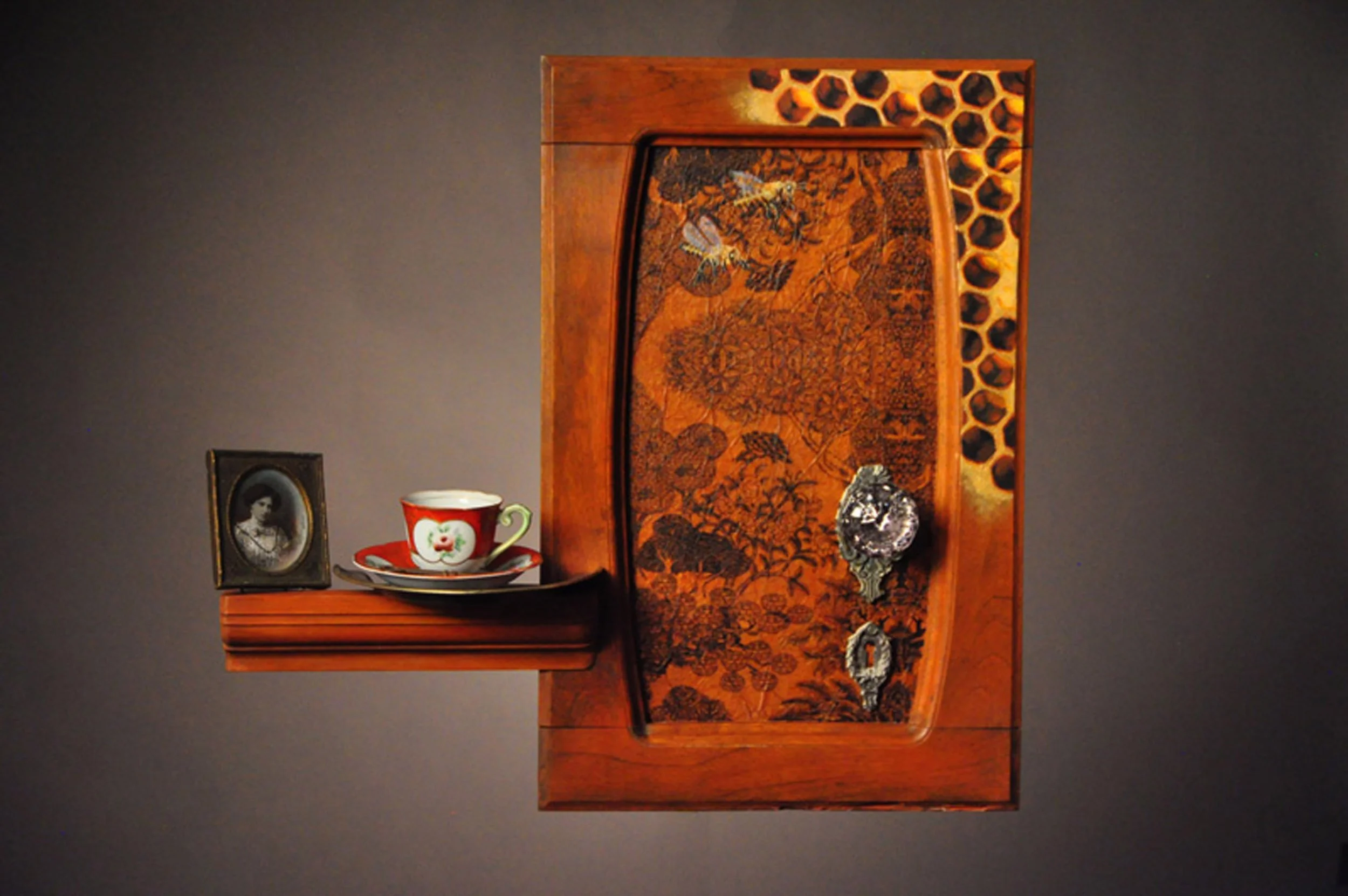 An antique wooden cabinet with decorative inlay, featuring a small portrait in a frame, a red teacup with a white floral design and matching saucer, and a decorative glass object.
