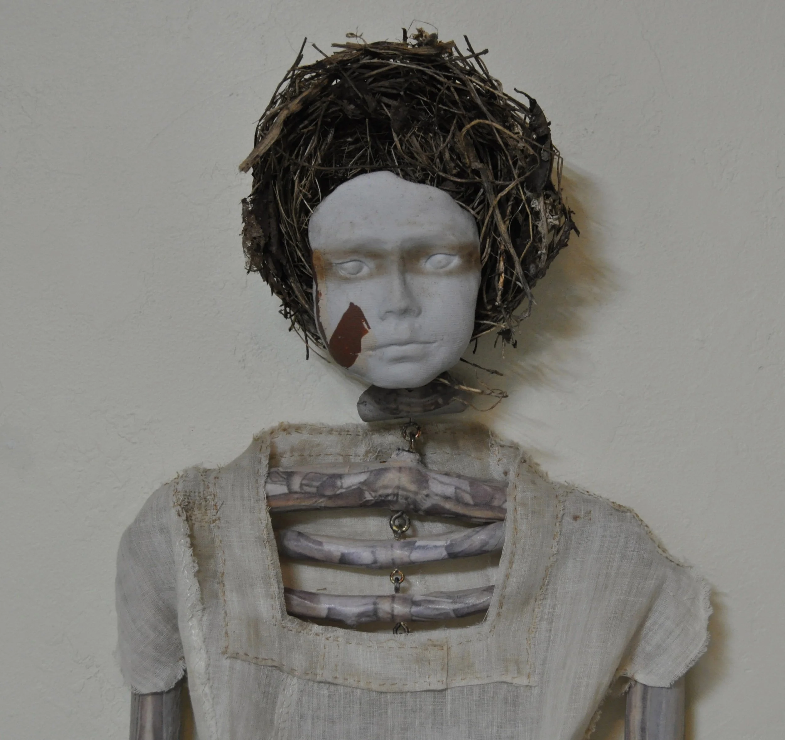 A white mannequin head with a face painting and a crown made of twigs, with a neutral background.