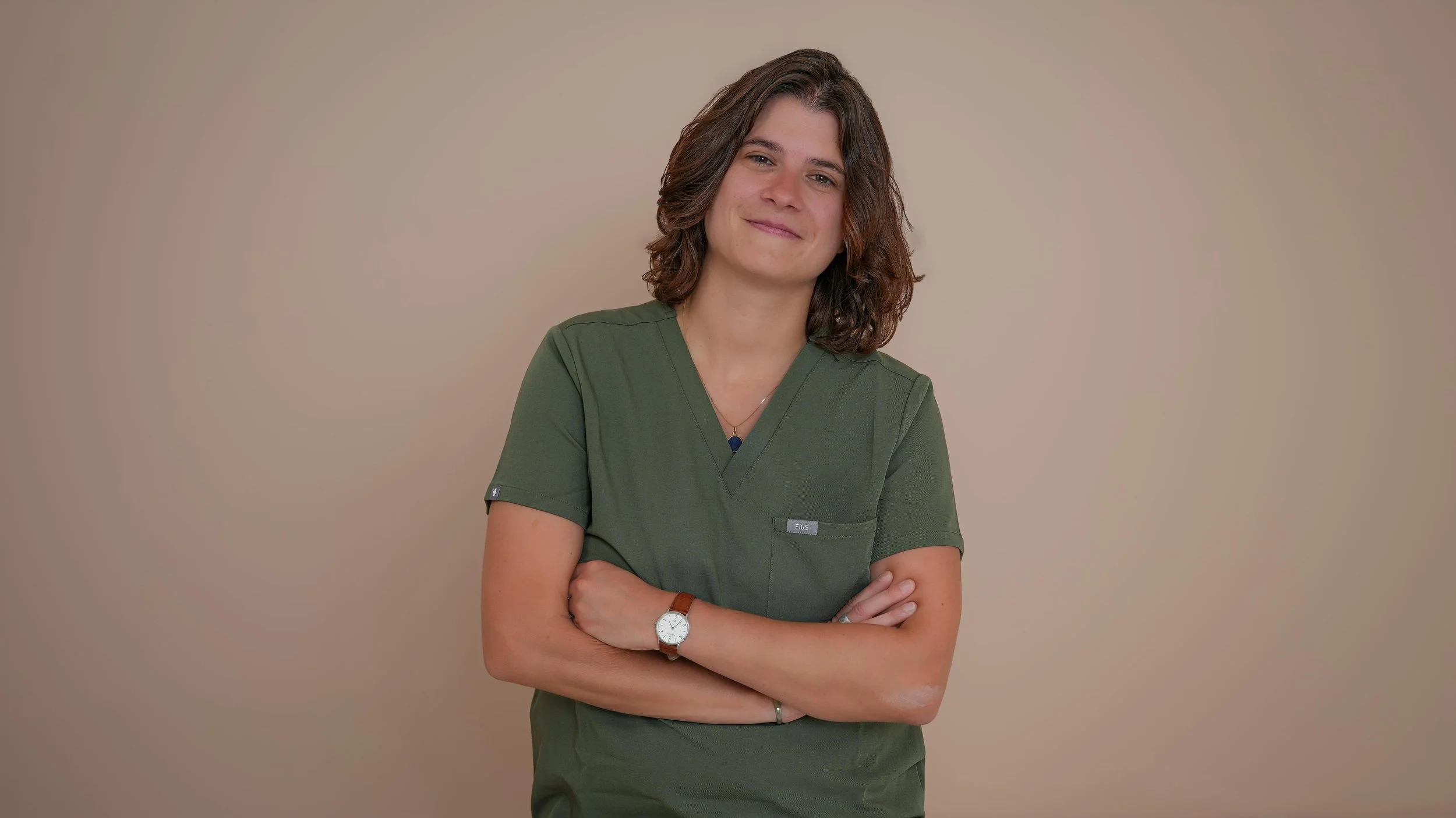 Une femme portant une blouse verte, souriant, avec les bras croisés, contre un fond beige.