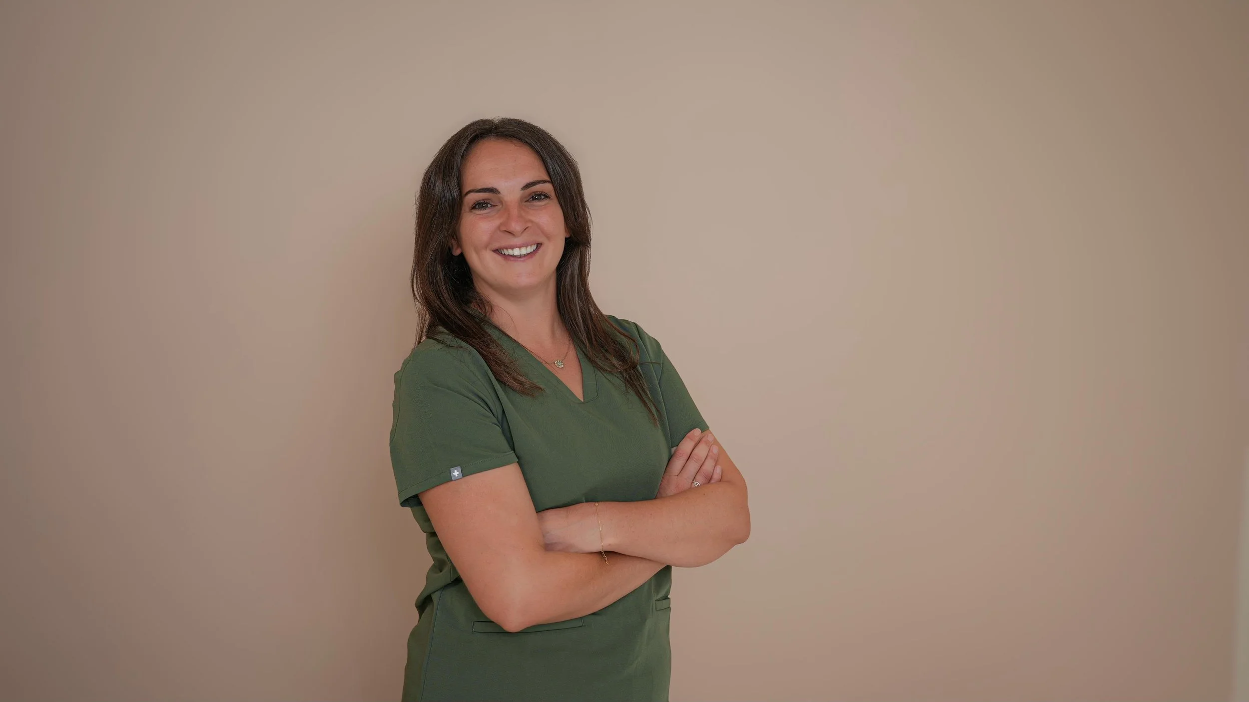 Une femme souriante, portant une tenue médicale verte, se tient contre un mur beige, avec les bras croisés.