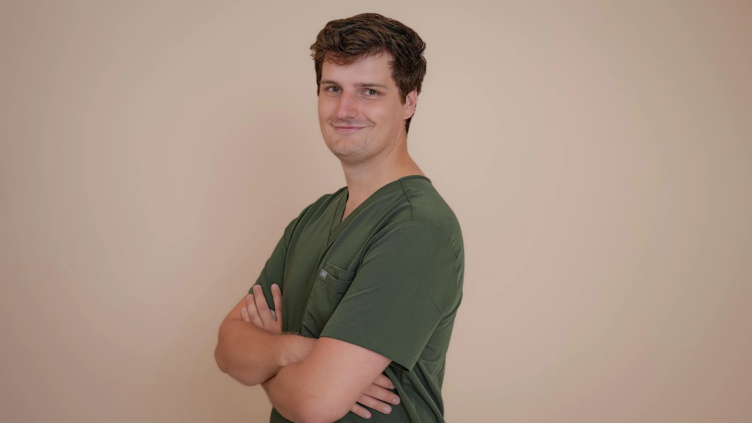 Jeune homme en tenue médicale avec bras croisés, souriant, fond neutre.