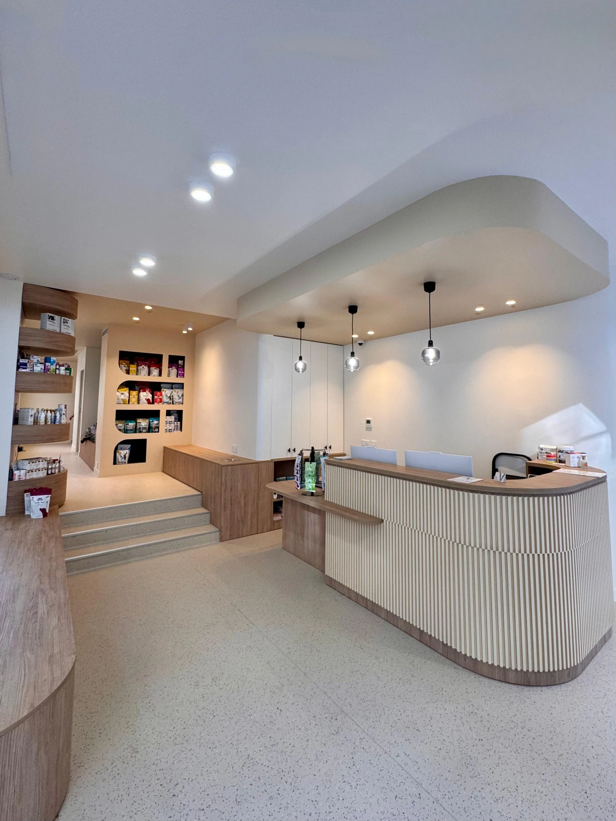Intérieur moderne avec comptoir en bois, suspension lumineuse, étagères avec produits, sol en PVC clair, murs blancs, ambiance lumineuse d'une clinique vétérinaire à Paris 15e