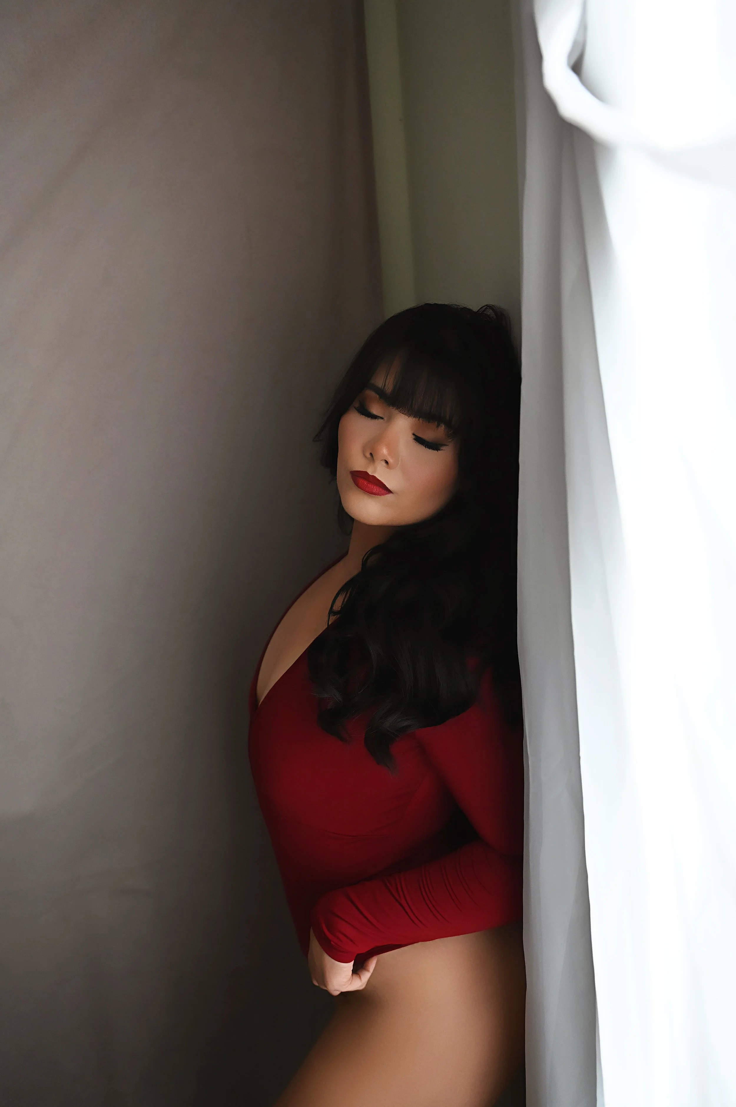 Woman in a red bodysuit posing by a window during her Shreveport boudoir session