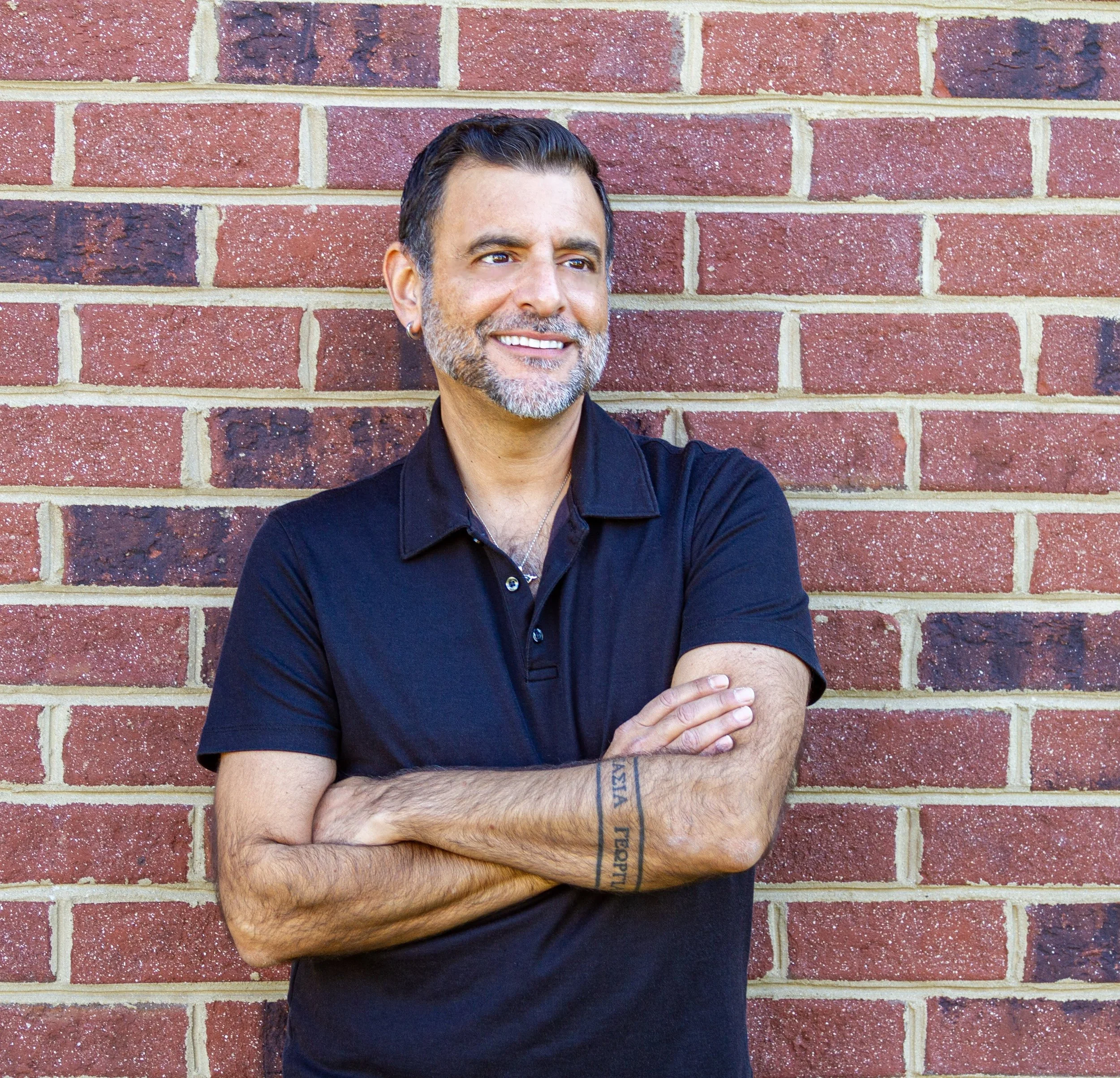 Close-up of a smiling man with short dark hair, facial hair, and earrings standing against a red brick wall with arms crossed, wearing a black shirt.
