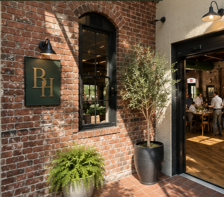 Exterior view of a restaurant featuring a brick wall, a black sign with gold letters 'BH', a large window, and an entrance door with people dining inside visible through the doorway.