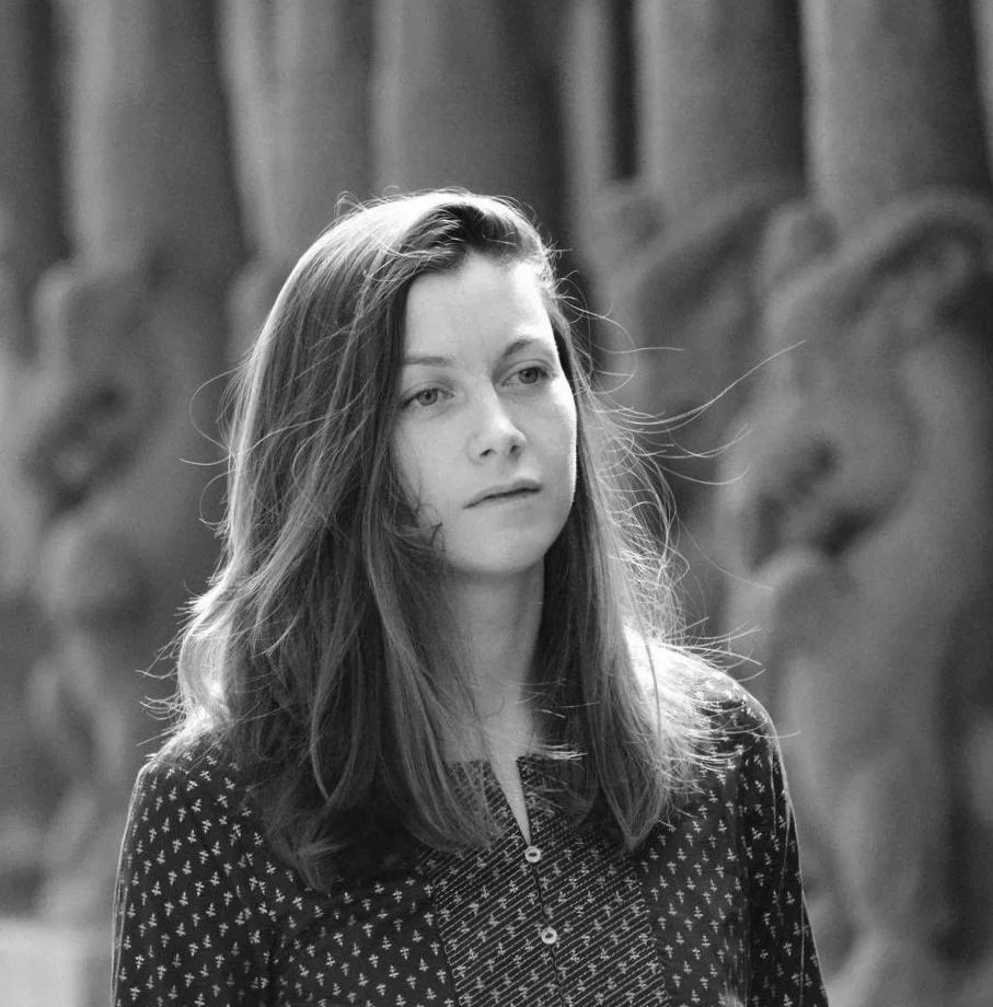 Black and white photo of a young woman with long hair, wearing a patterned top, standing in front of a blurred background.