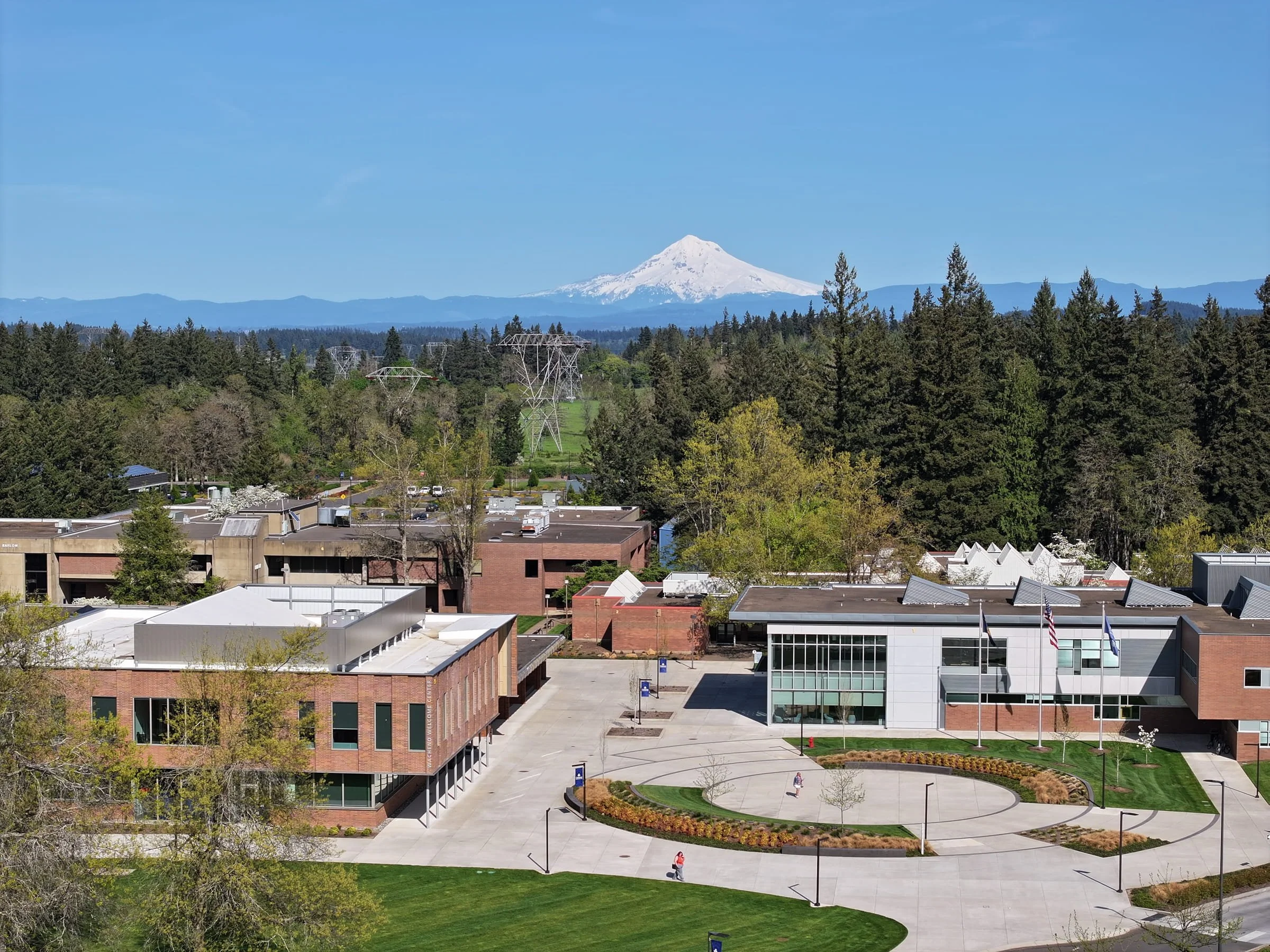 A university campus with modern buildings, a circular paved area with a small garden, and a green lawn, with Mount Hood in the background under a clear blue sky.