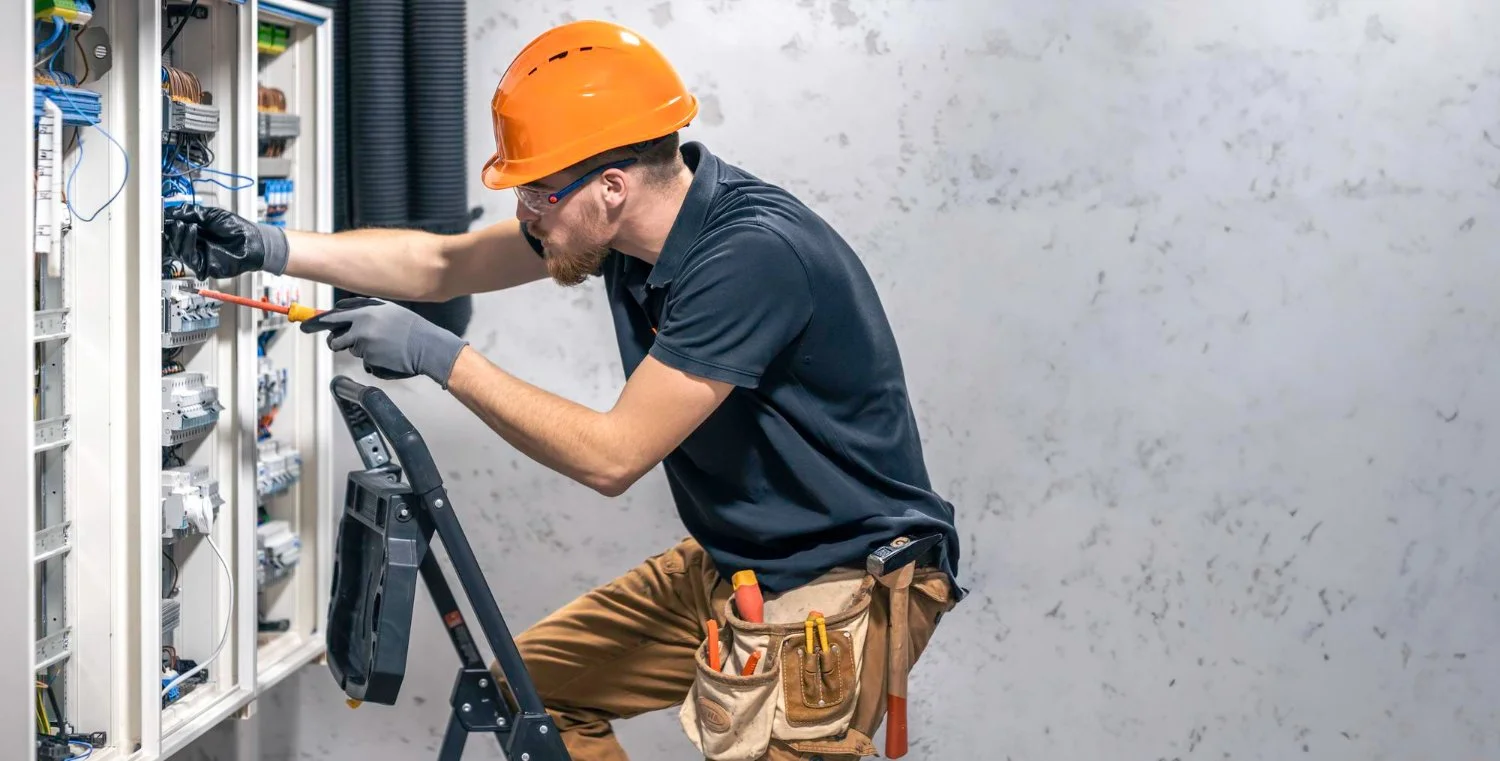 Technician working on electrical panel, wearing orange safety helmet, using screwdriver, with tools in pouch, standing on ladder.