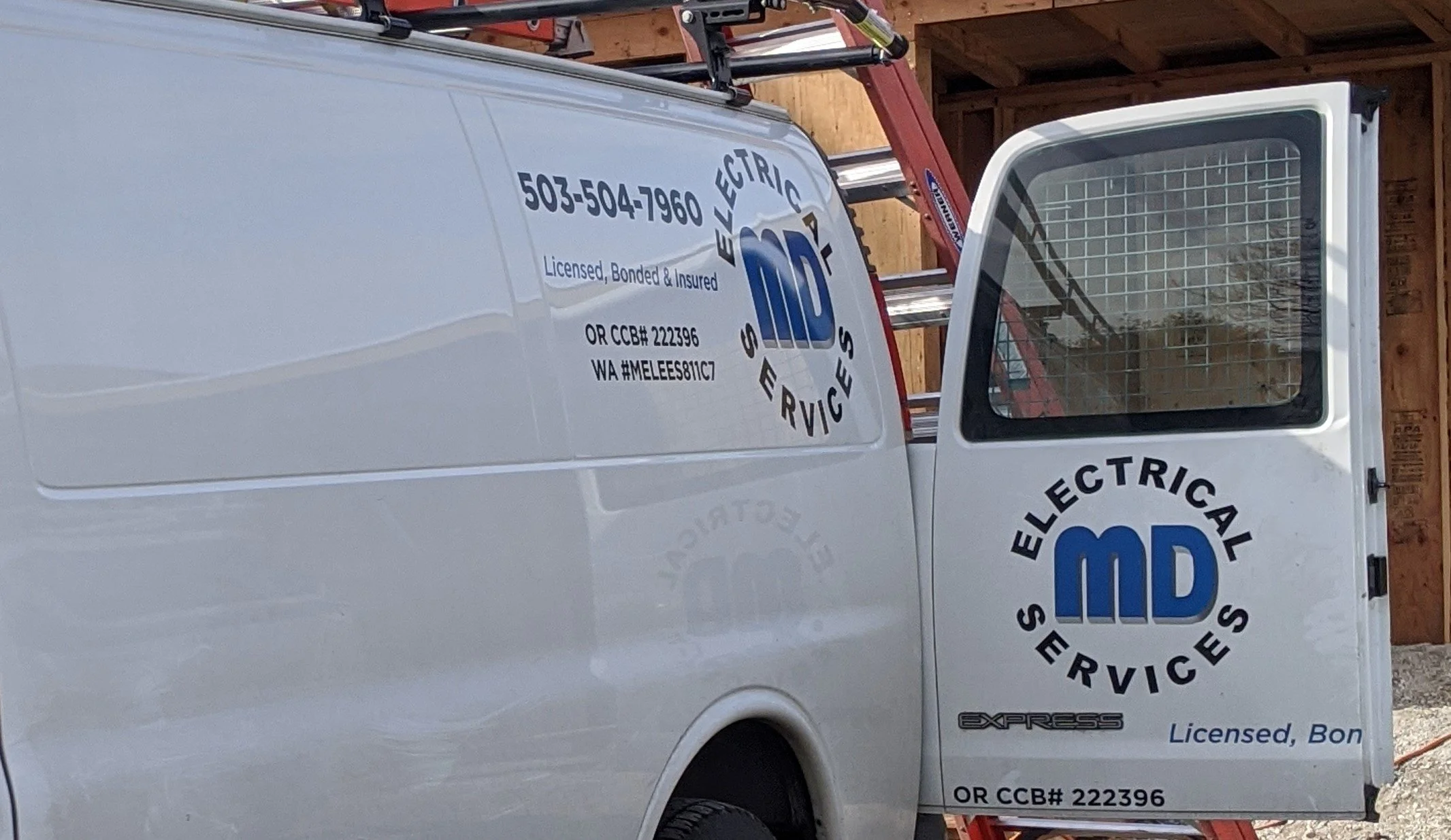 A white van with logo and contact information for an electrical services company, parked in front of a wooden structure under construction.