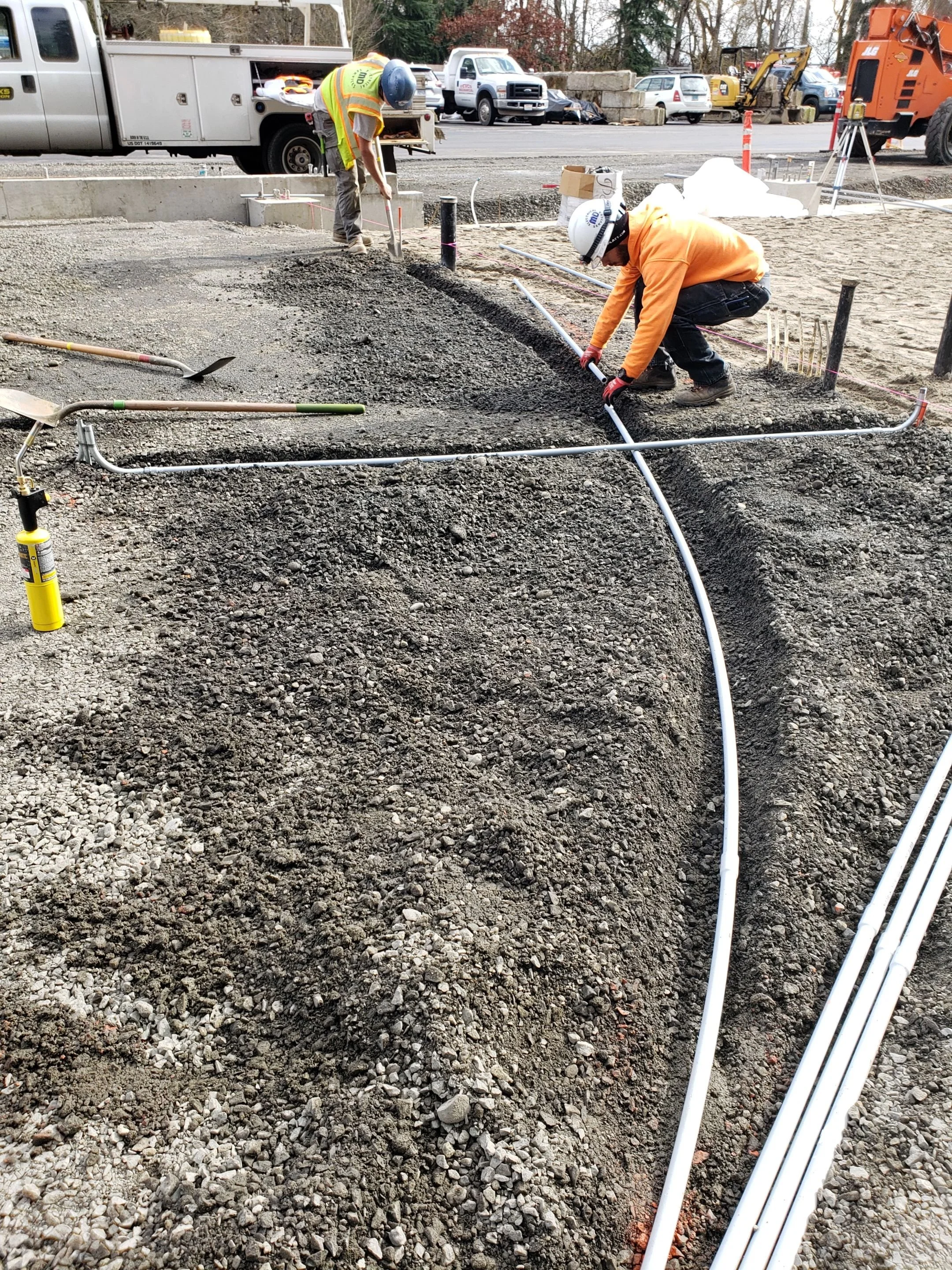 Construction workers installing conduit pipes at a construction site, with trucks and construction equipment in the background.