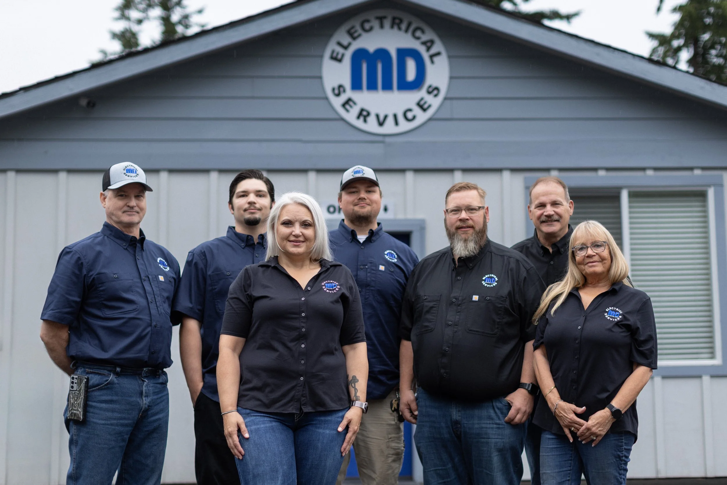 Group of six people standing outside in front of a building with a sign that reads 'Electrical Services' and a large logo with the initials 'MD'.