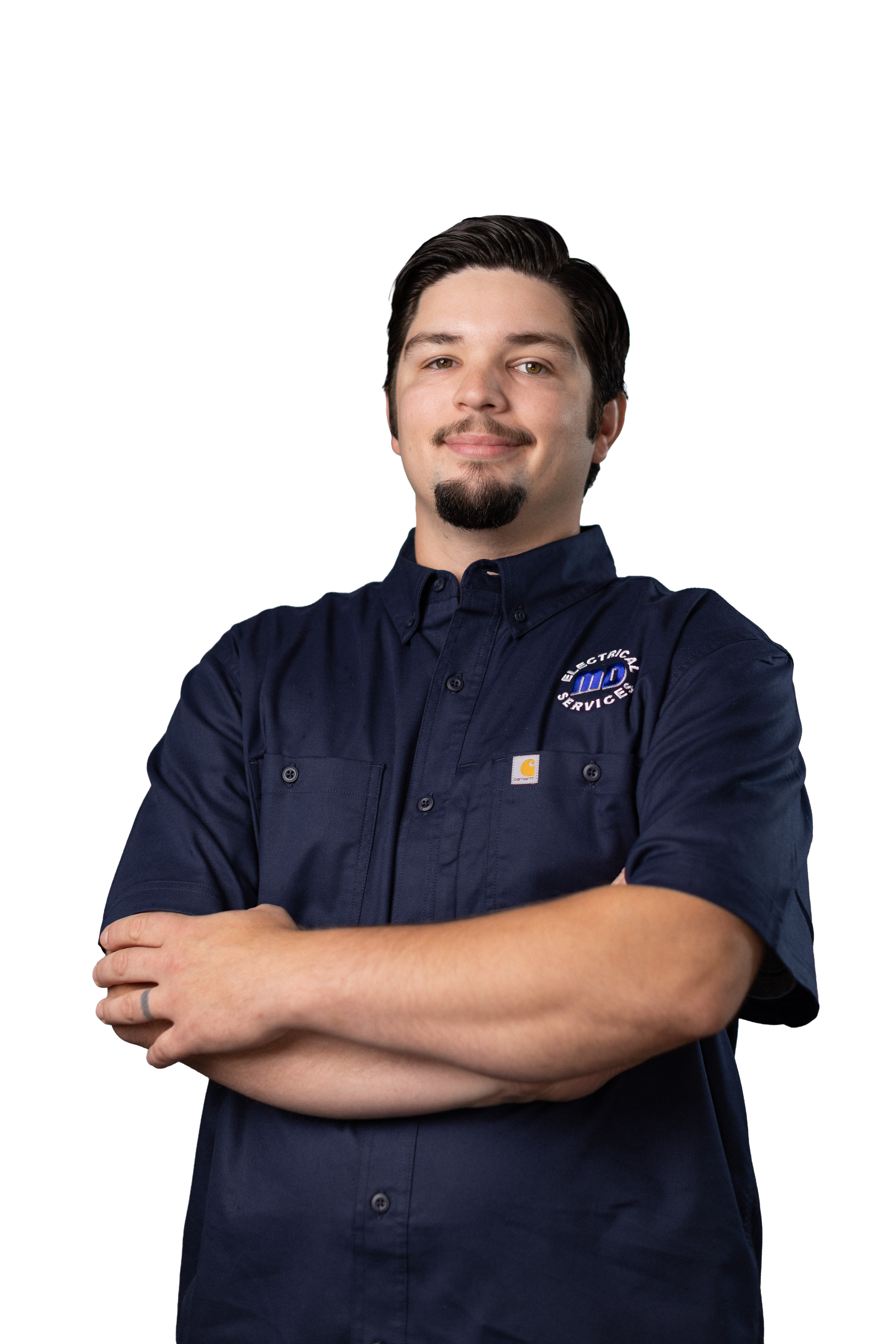A young man with dark hair, mustache, and goatee, smiling with arms crossed, wearing a navy blue work shirt with company logos, standing against a plain background.