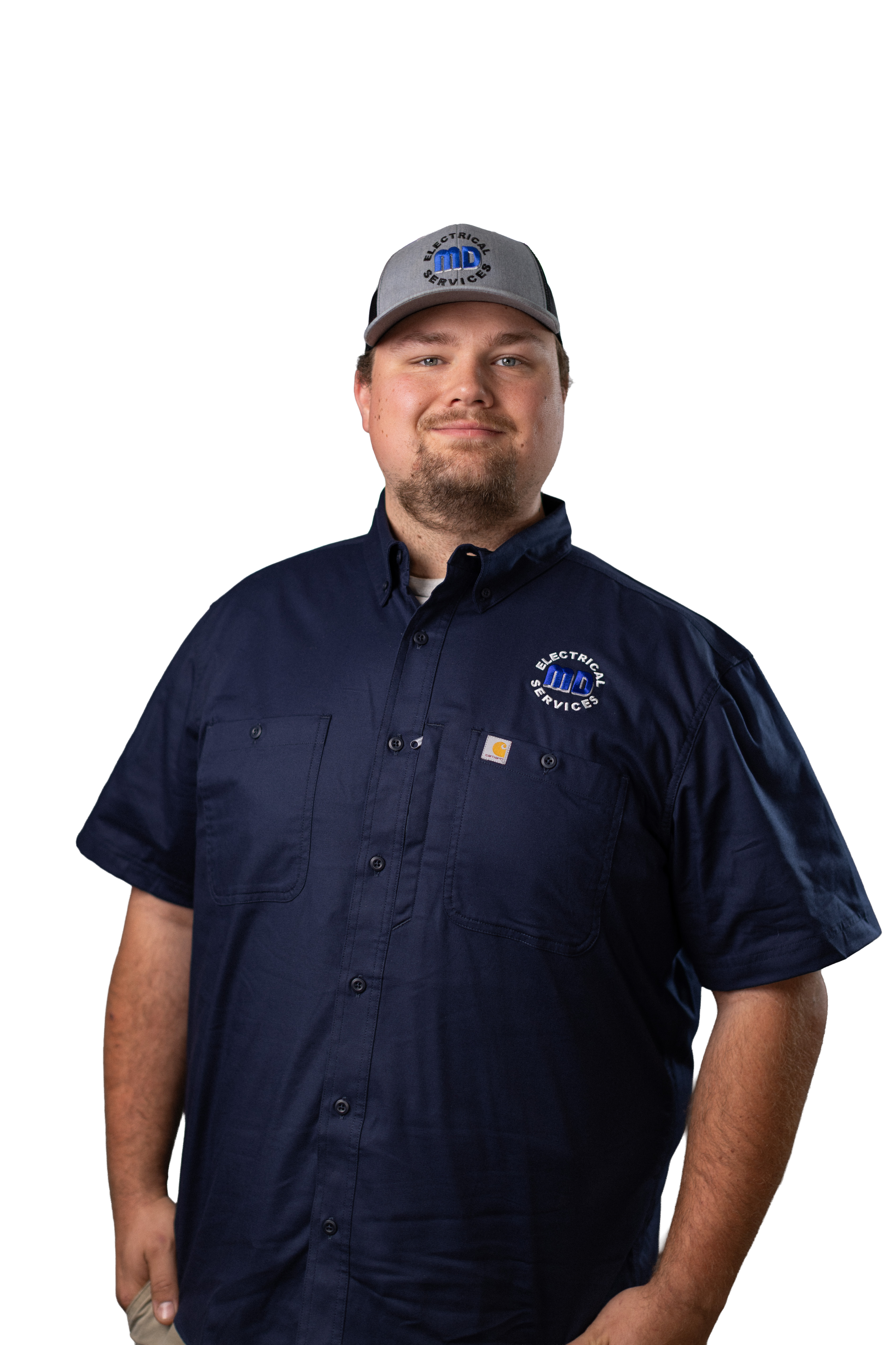 A young man with light skin, short brown hair, and a goatee, smiling, wearing a gray and black baseball cap with a logo that reads 'Electrical Services' and a dark blue work shirt with the same logo embroidered on the left chest.