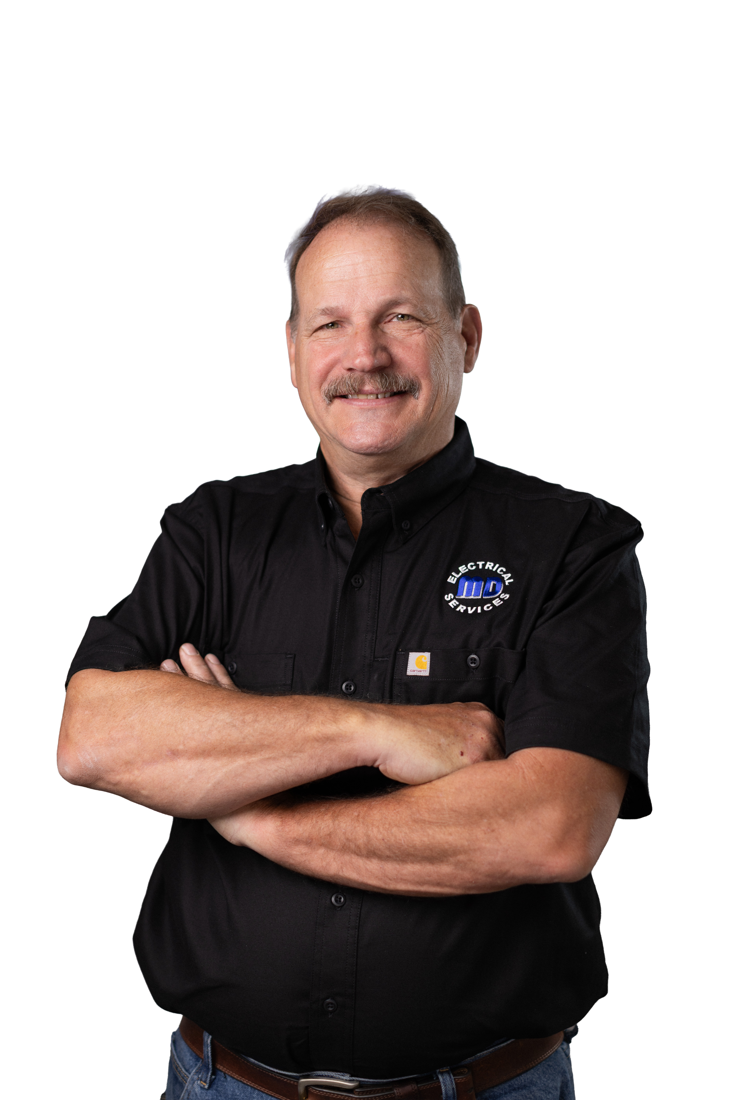A man with a mustache and beard, smiling, standing with arms crossed, wearing a black collared shirt with a company logo that reads 'Electrical Services MD,' blue jeans, and a brown belt.
