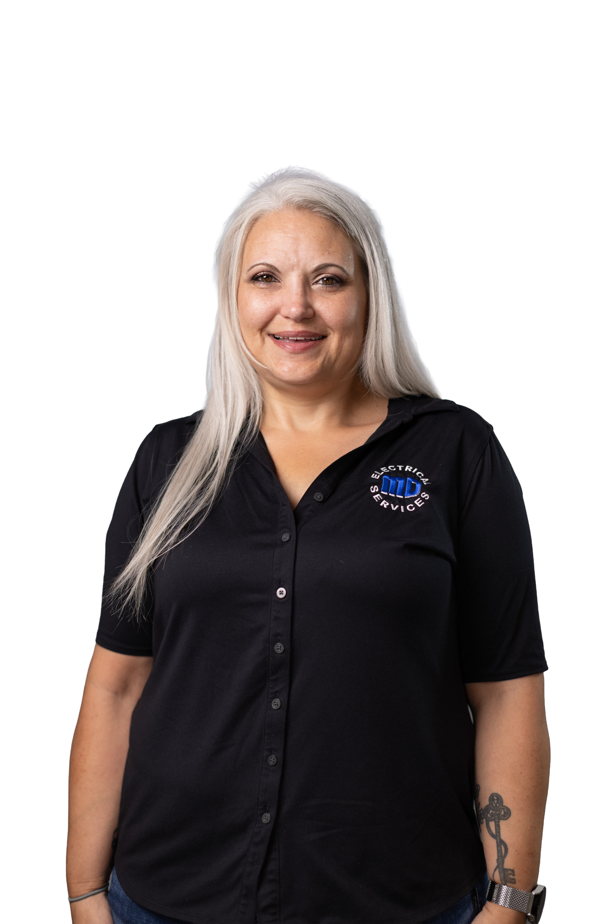 A woman with long platinum blonde hair, smiling, wearing a black collared shirt with a logo that reads 'ELECTRICAL SERVICES'. She has a tattoo of a cross on her left forearm and wears a silver watch on her left wrist, standing against a plain background.
