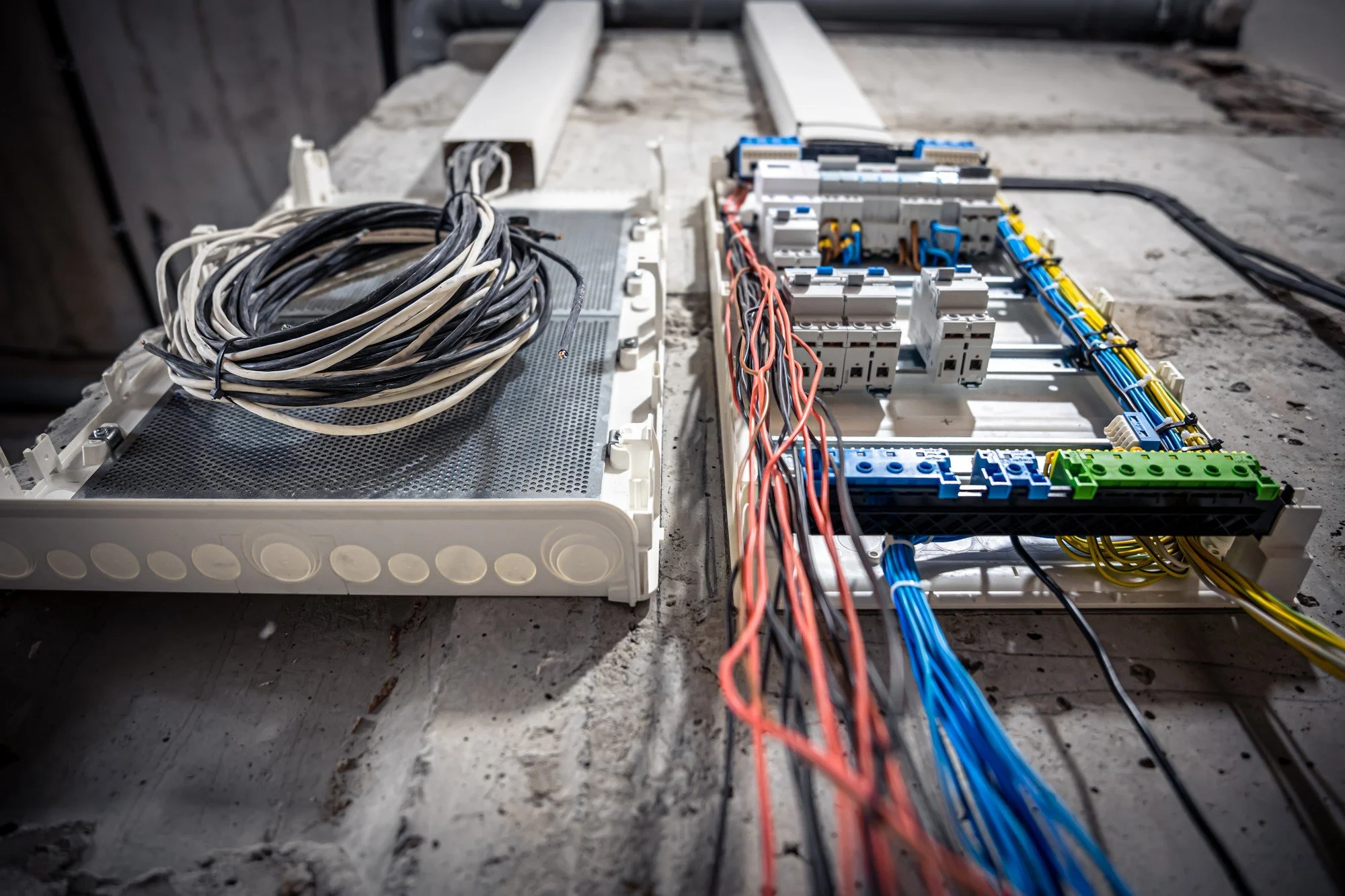 Open electrical box with bundled black and white wires, and organized red, blue, yellow, and green wires on a concrete surface.