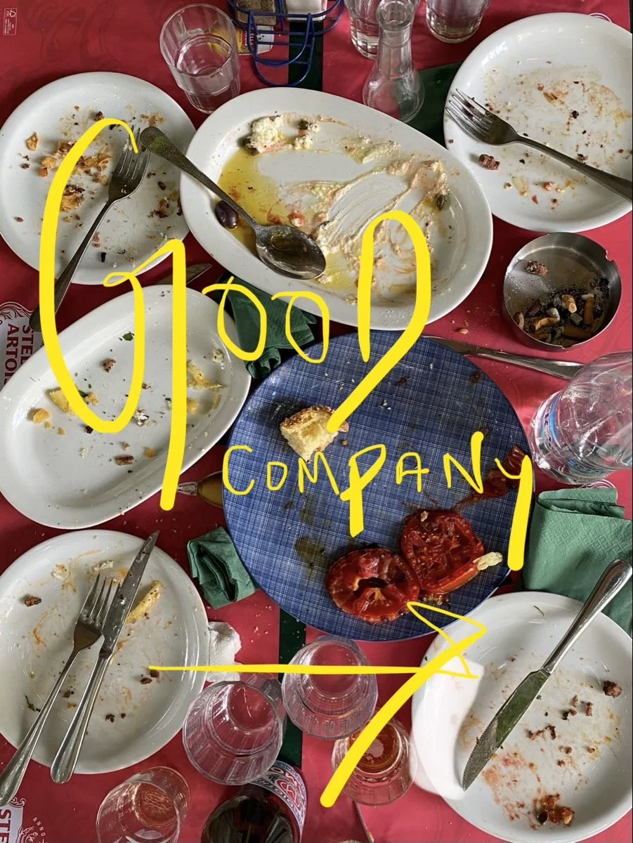 A table with empty and partially eaten plates, glasses, and utensils, with a few remaining slices of tomato and some food scraps. The table appears to be at the end of a meal, with a red tablecloth and some green napkins, and handwritten yellow text on the image reads 'happy company'.