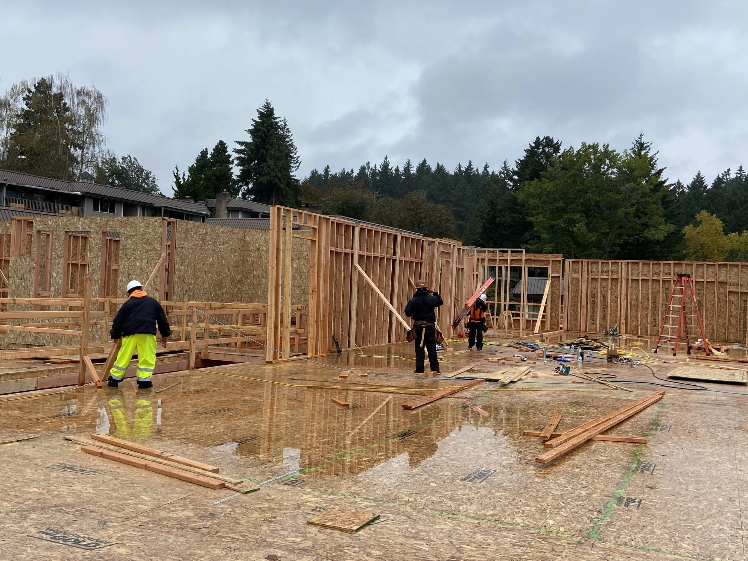 Construction site with wooden framing for a building and workers wearing safety helmets and reflective clothing. Overcast sky, trees in the background.