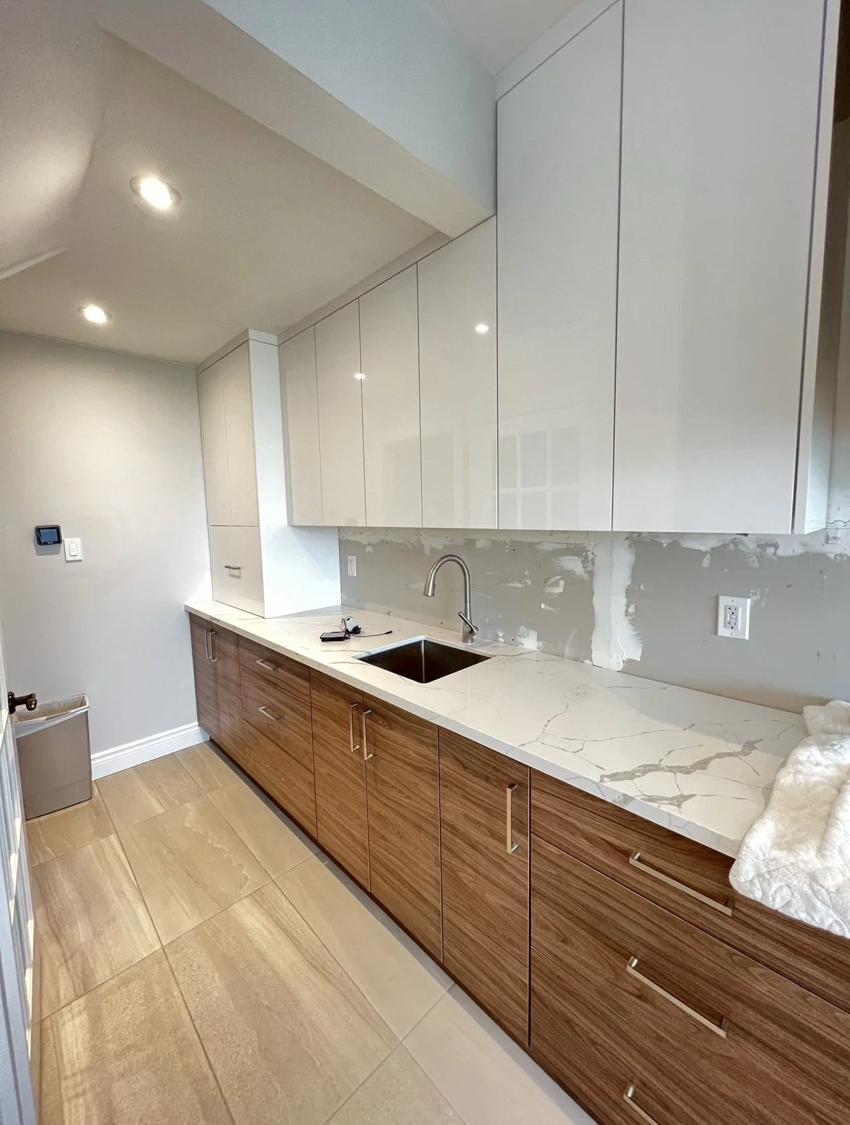 Kitchen with wood lower cabinets, white upper cabinets, marble countertop, stainless steel sink, and beige tiled floor, in a room under renovation.