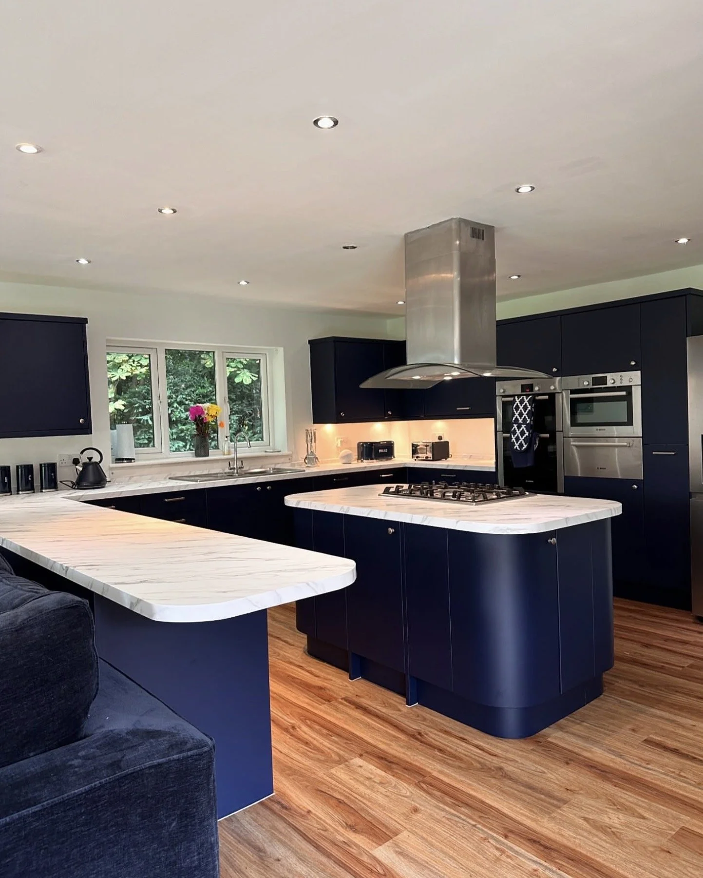 Modern kitchen with dark navy cabinets, white marble countertops, and a central island with a gas stove. A window with a vase of colorful flowers above the sink. Recessed lighting in the ceiling, and hardwood floors.