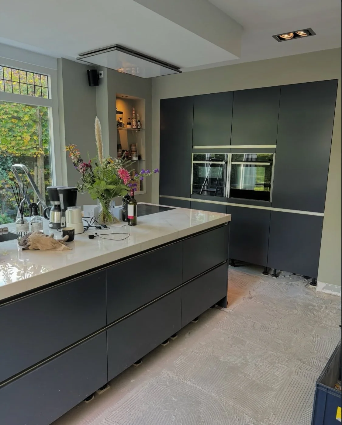 Modern kitchen with dark cabinetry, white island countertop, and a vase of flowers on the counter. There is a large window with outside greenery and various small appliances and items on the counter.