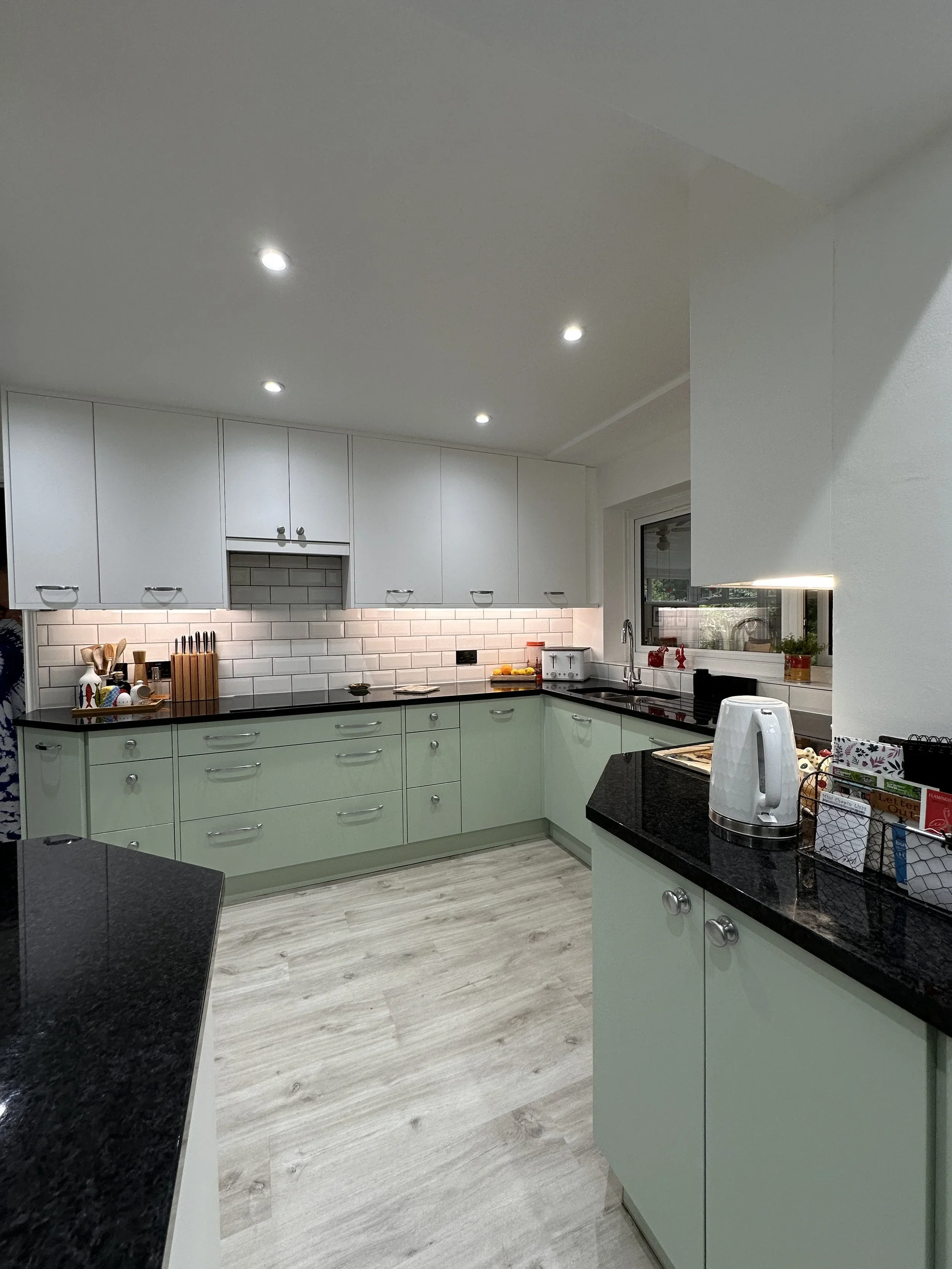Modern kitchen with light green lower cabinets and white upper cabinets, black granite countertops, white subway tile backsplash, and various small appliances and utensils on the counters, illuminated by recessed ceiling lights and a window.