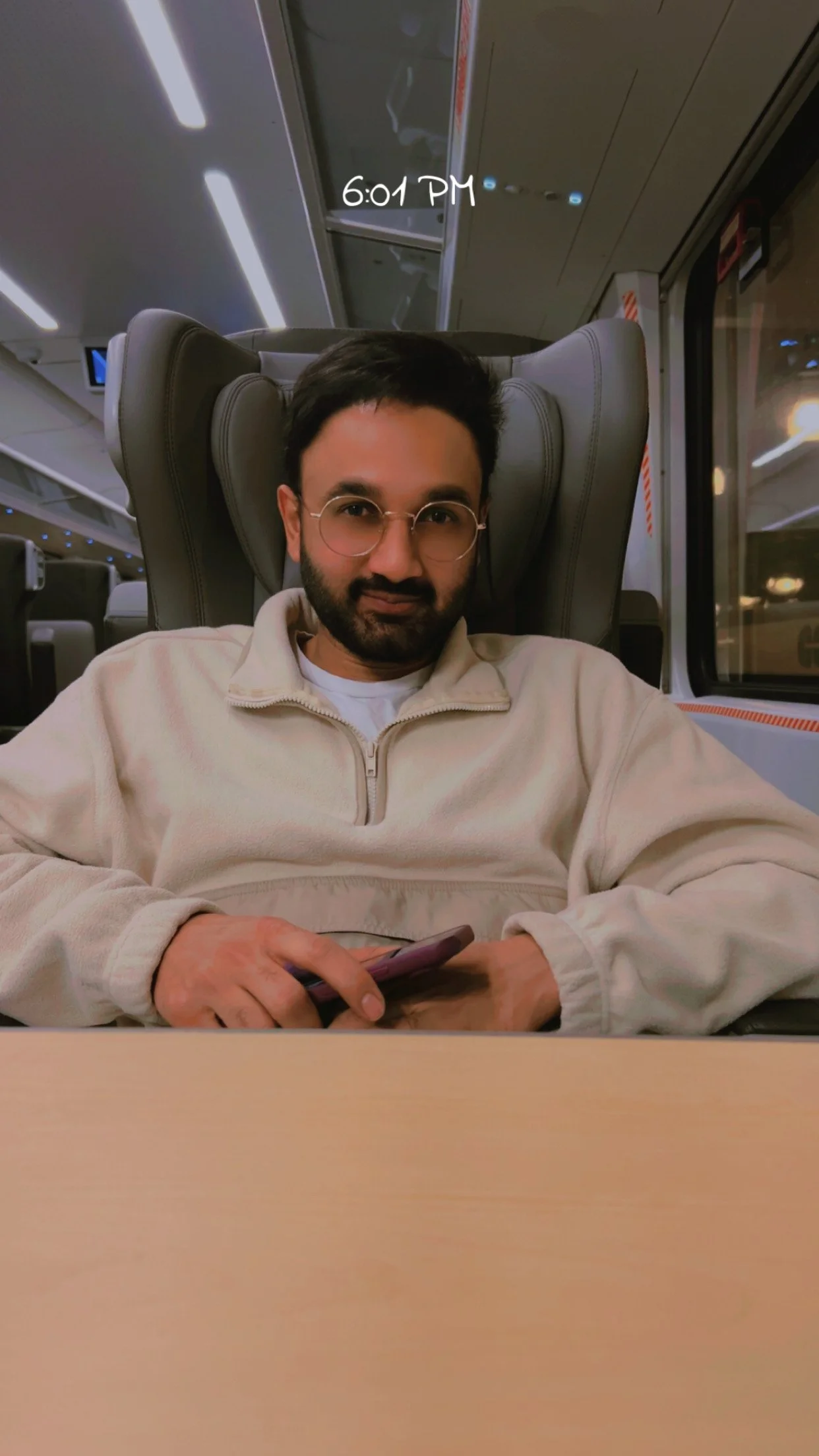 A man with glasses and a beard wearing a beige pullover, sitting on a train, holding a smartphone, with a window showing the night outside; a sign on the window indicates the time is 6:01 PM.