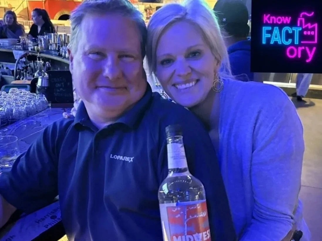 A man and woman sitting close together at a bar or restaurant, smiling at the camera. The man holds a bottle of alcohol, and there are glasses and bottles on the table. Neon sign in background reads 'Know FACT ory'.