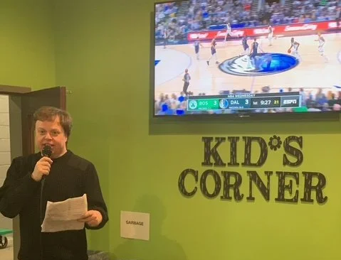 A young man with a microphone and paper standing in front of a green wall . A large TV screen displaying a basketball game is mounted on the wall. The wall also has a sign that reads 'KID'S CORNER'.