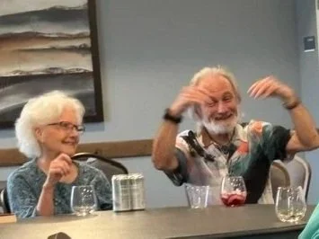 Two older adults, a woman and a man, sit at a table with glasses of water and a glass of red drink, smiling and enjoying a cheerful moment.