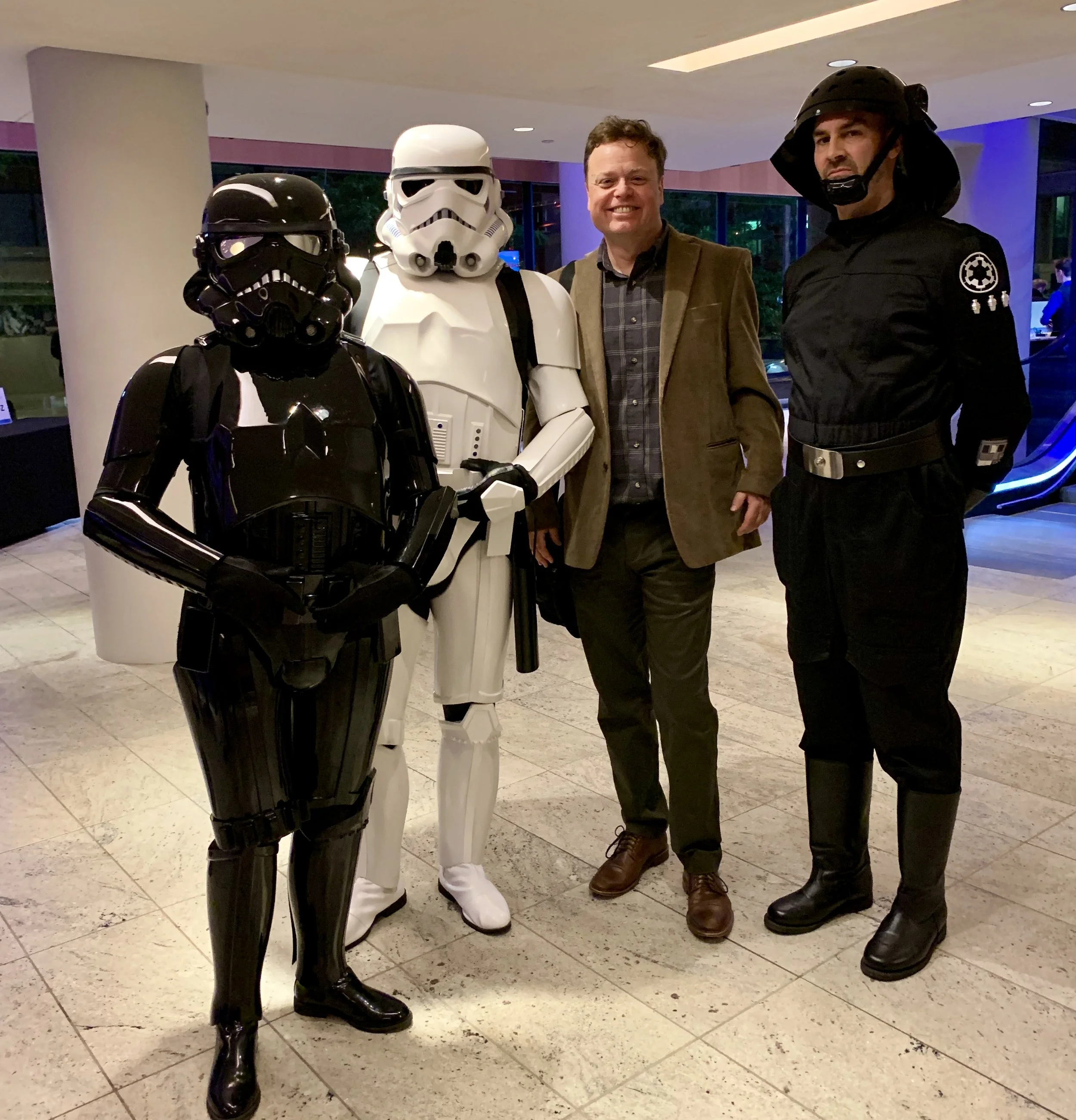 Group of four people, three dressed as Star Wars characters in costume and one man in casual clothing, standing indoors with large windows in the background.