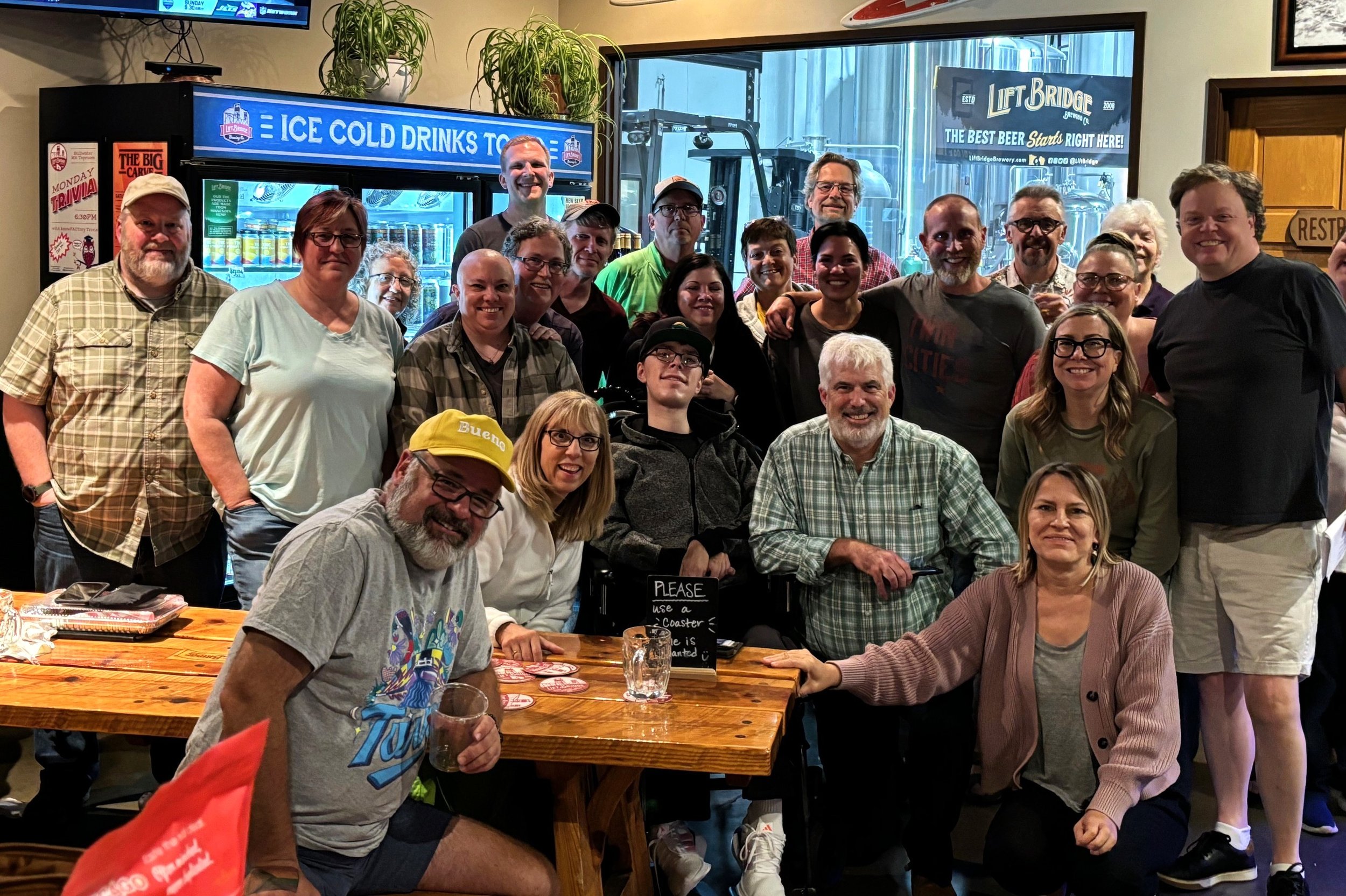 A group of about 20 people gathered inside a restaurant or pub, smiling for a group photo. Some are standing behind a wooden table, and others are seated. There are promotional signs and beer branding in the background, including a sign for ice cold drinks and a brewery sign. The atmosphere appears lively and friendly.