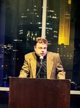 A man in a blazer speaking at a podium, with a city skyline visible through large windows behind him.