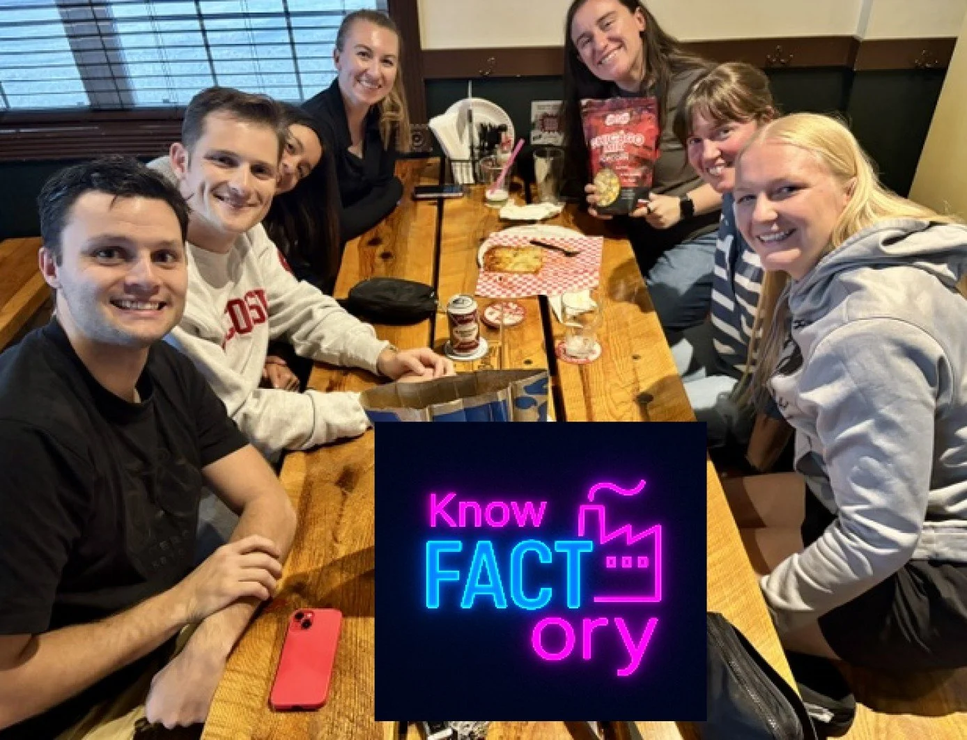 A group of seven friends sitting around a wooden table inside a restaurant, smiling at the camera with snacks and drinks, and a neon sign reading 'Know FACT ory' placed in front of the table.