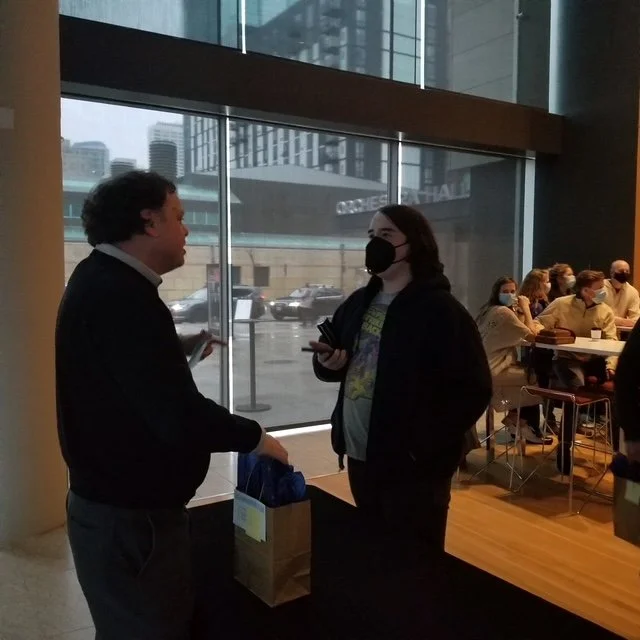 Two people talking inside a modern office or conference area, with several seated individuals wearing masks in the background. Large windows reveal a cityscape with tall buildings.