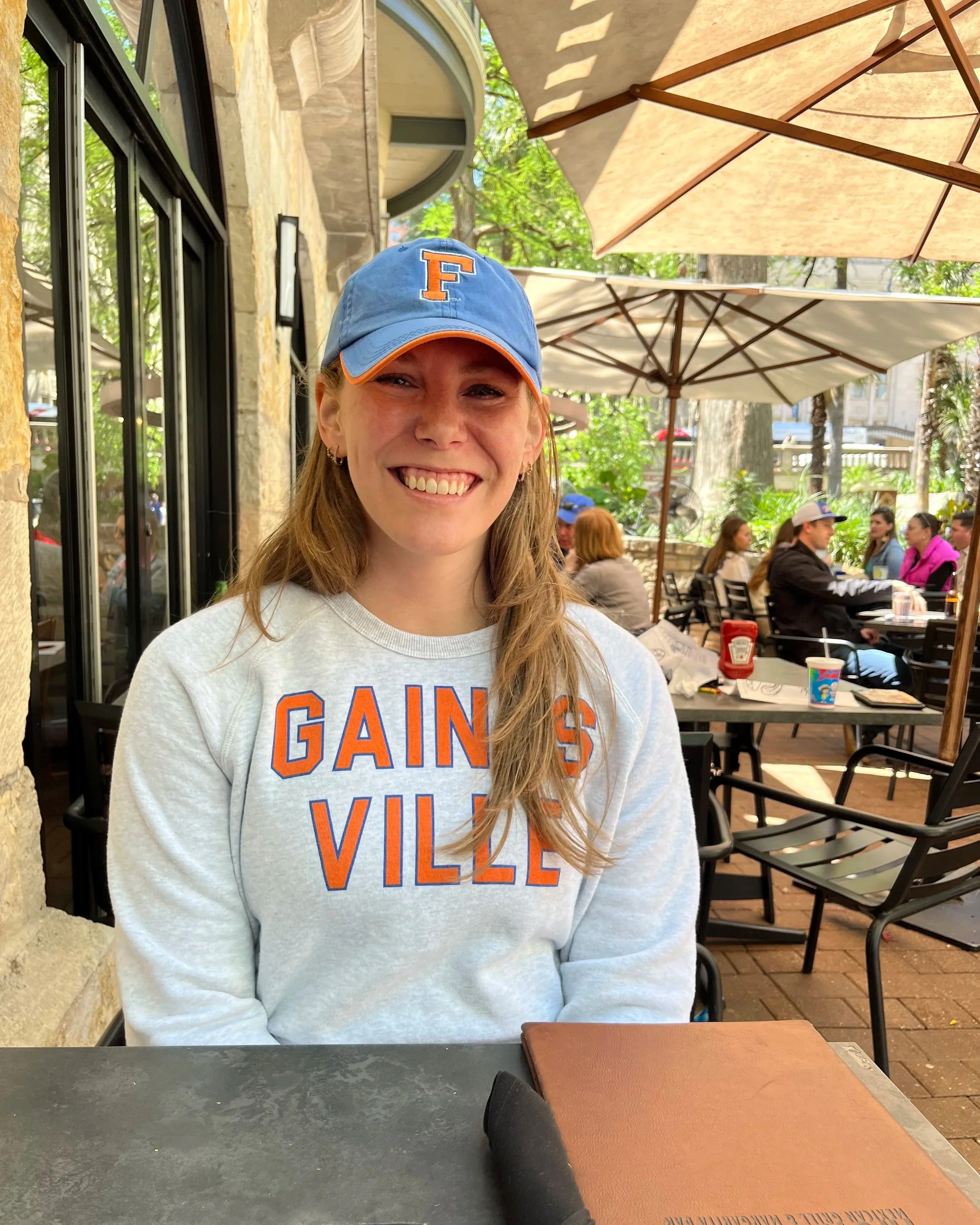 A young woman with long blonde hair wearing a light gray sweatshirt and a blue cap with an orange letter 'F' in a restaurant patio dining area with umbrellas, trees, and other patrons in the background.
