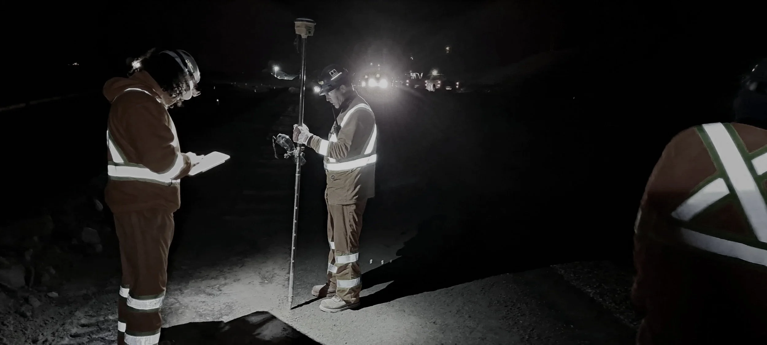 Nighttime scene of construction workers inspecting a road, with one holding a measuring pole and another taking notes, illuminated by headlights in the background.