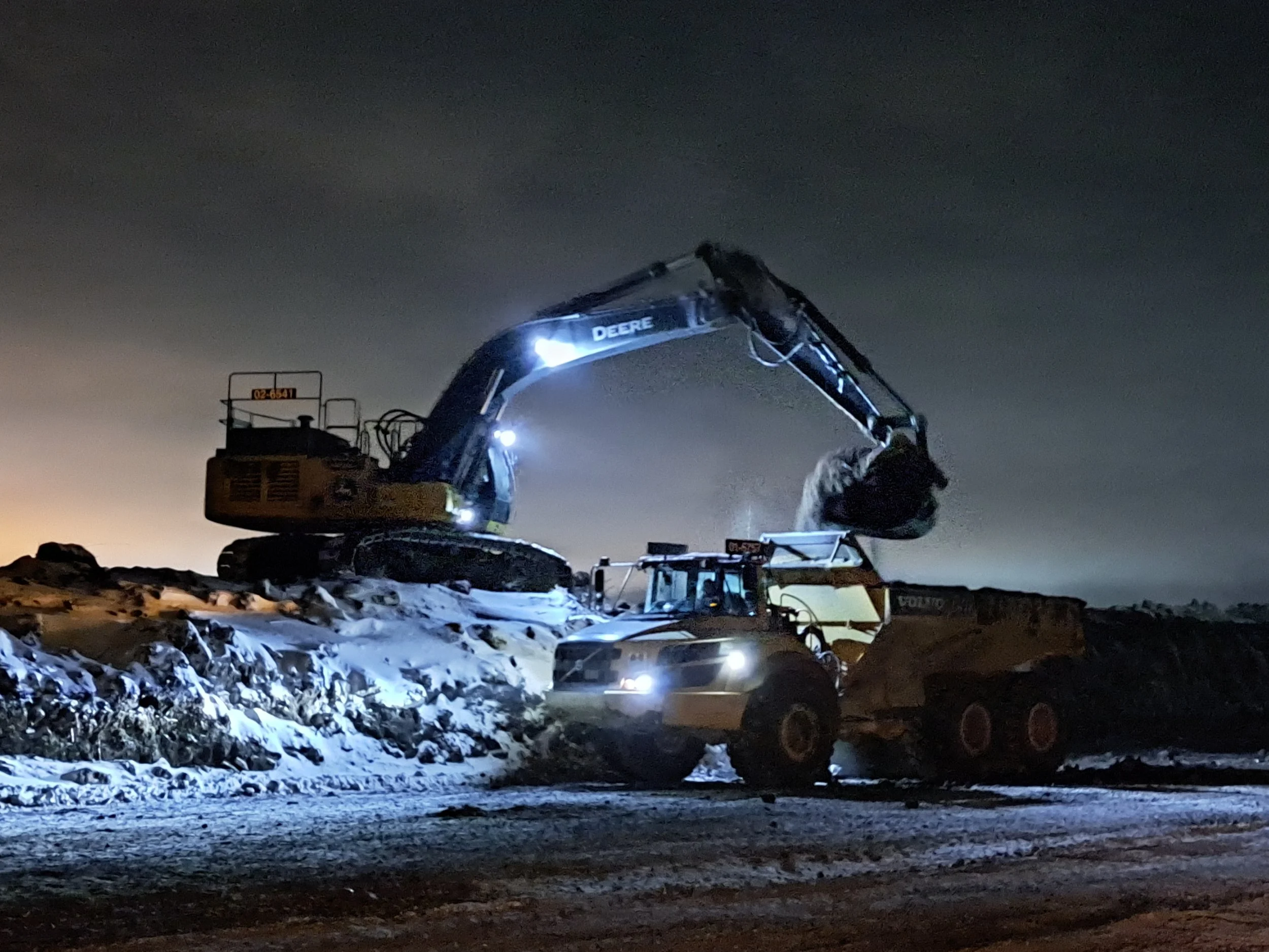 A construction excavator working during nighttime, illuminated by bright lights, with a snowy ground and dark cloudy sky.